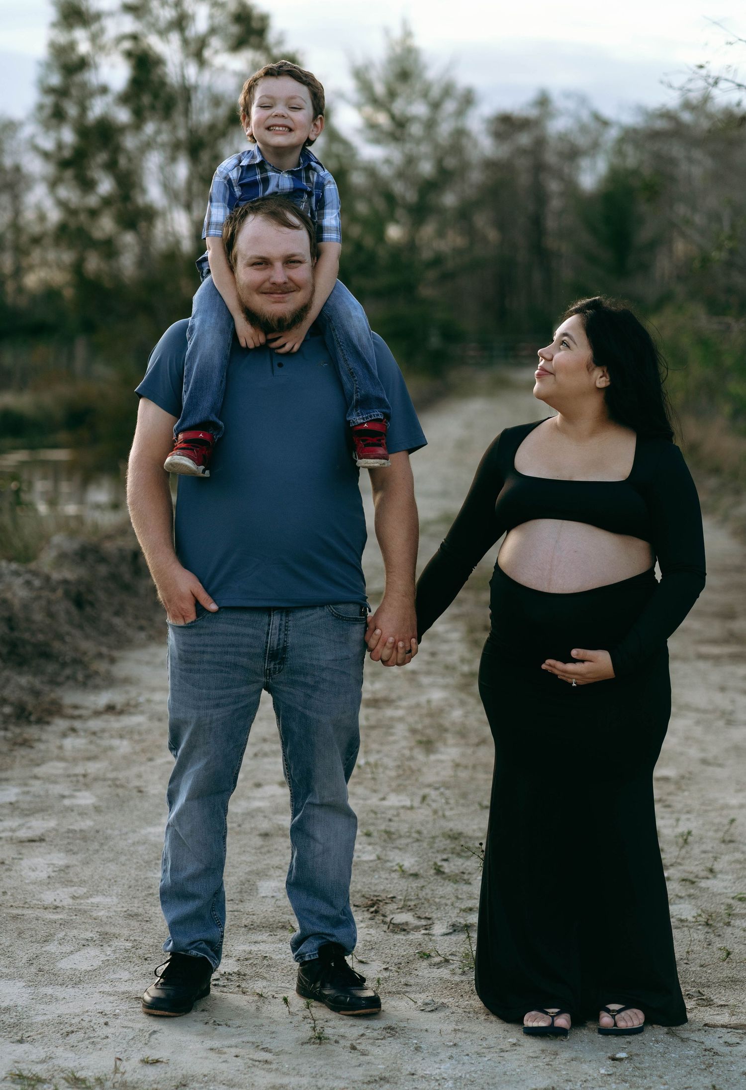 A family maternity photo outdoors with a pregnant woman in black and a child sitting on shoulders.