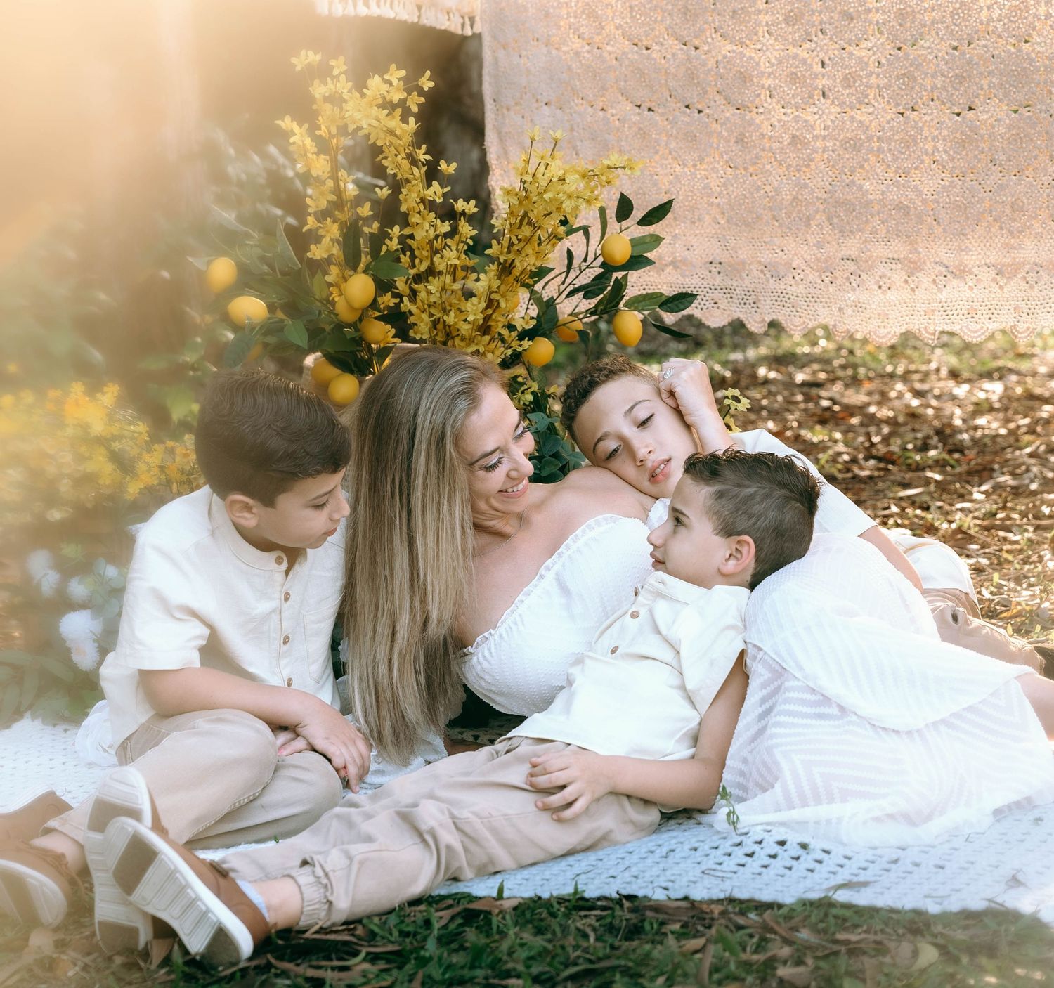 Family lying on white blanket outdoors with golden sunlight filtering through trees in autumn setting.