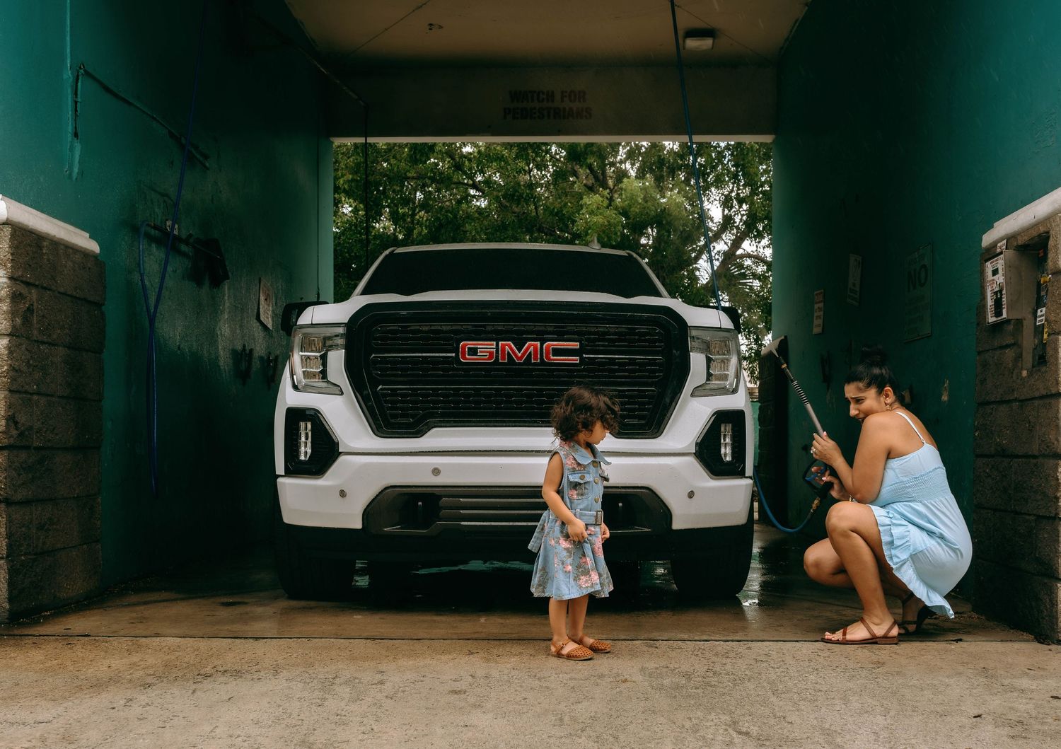Woman and child washing a white GMC truck in a garage with a pressure washer on a sunny day.