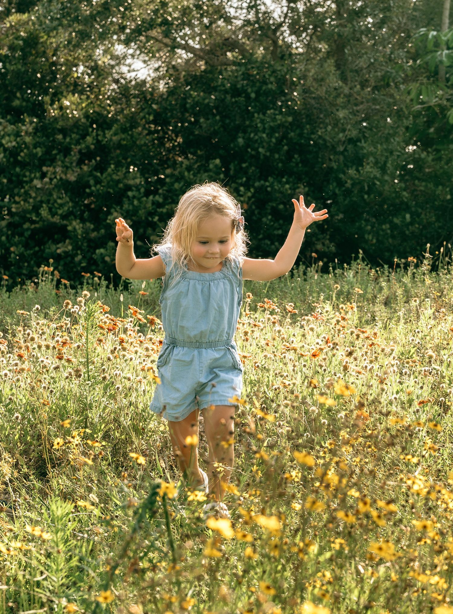 A joyful blonde toddler girl in a blue romper runs through a sunny wildflower meadow with arms raised.