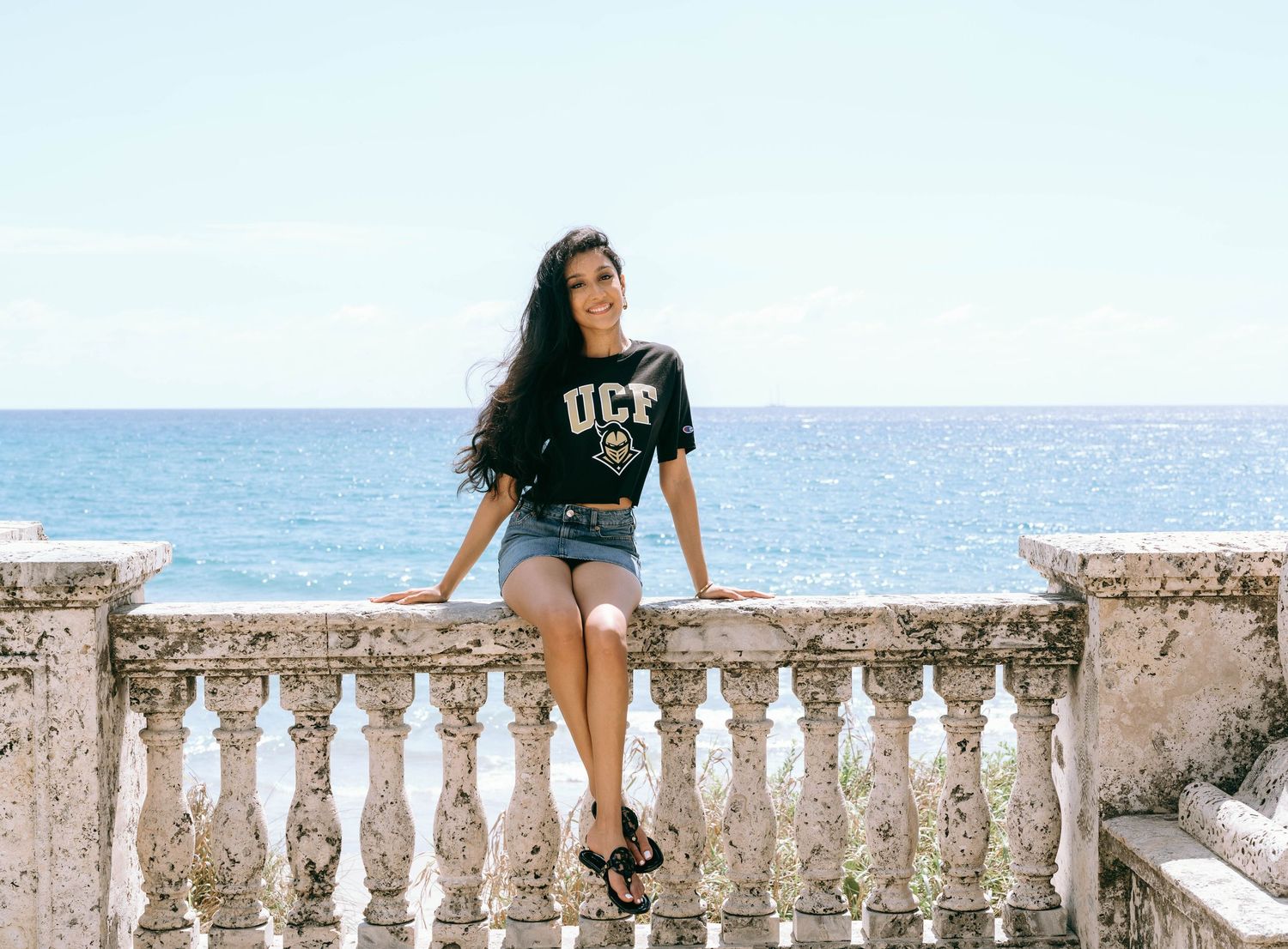 Young woman in UCF shirt sitting on stone balustrade with ocean view behind her on Worth Avenue Palm beach.
