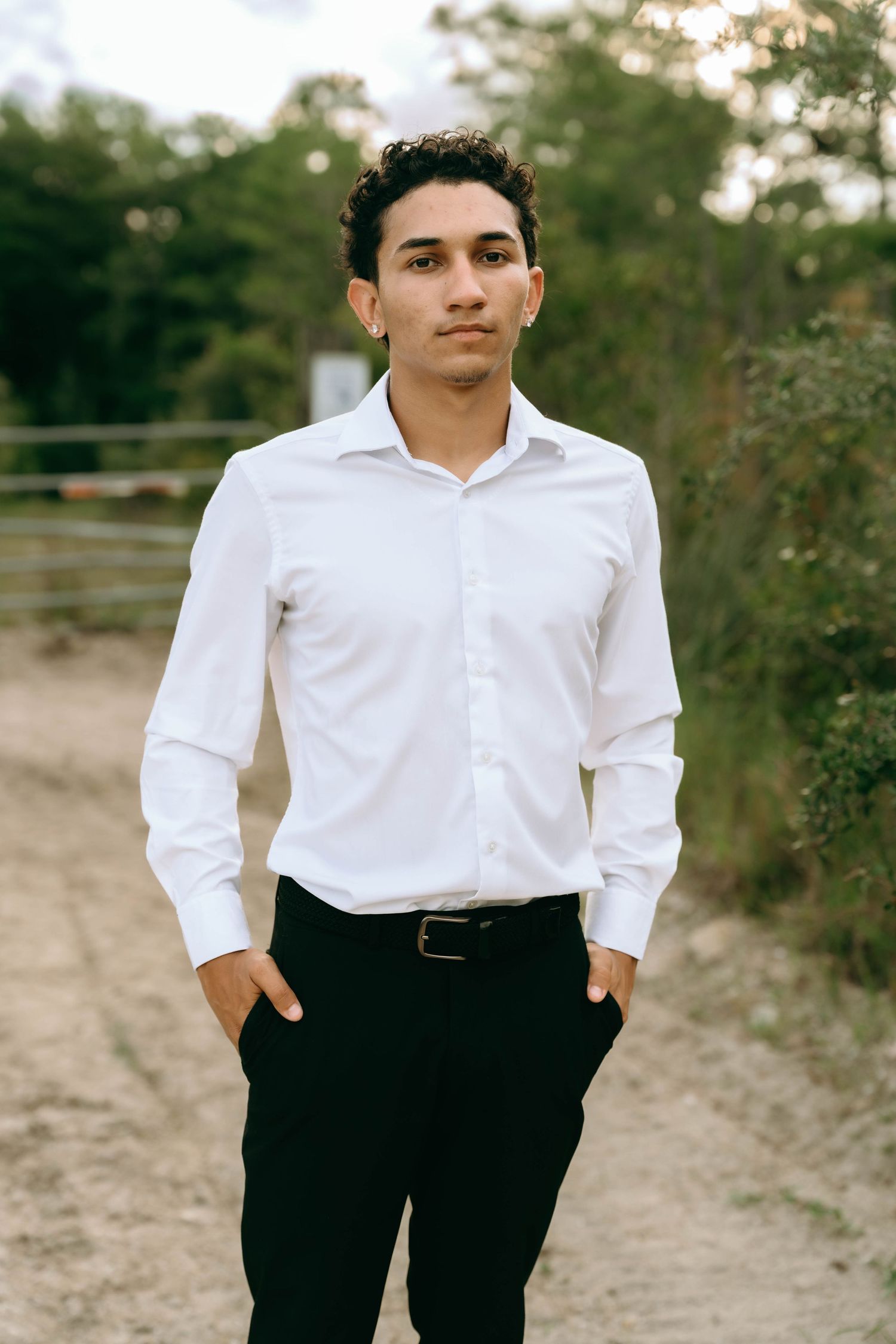 Young man in white dress shirt and black pants stands on a dirt path surrounded by trees. Senior Photos at Colt Farms in Boynton Beach, FL