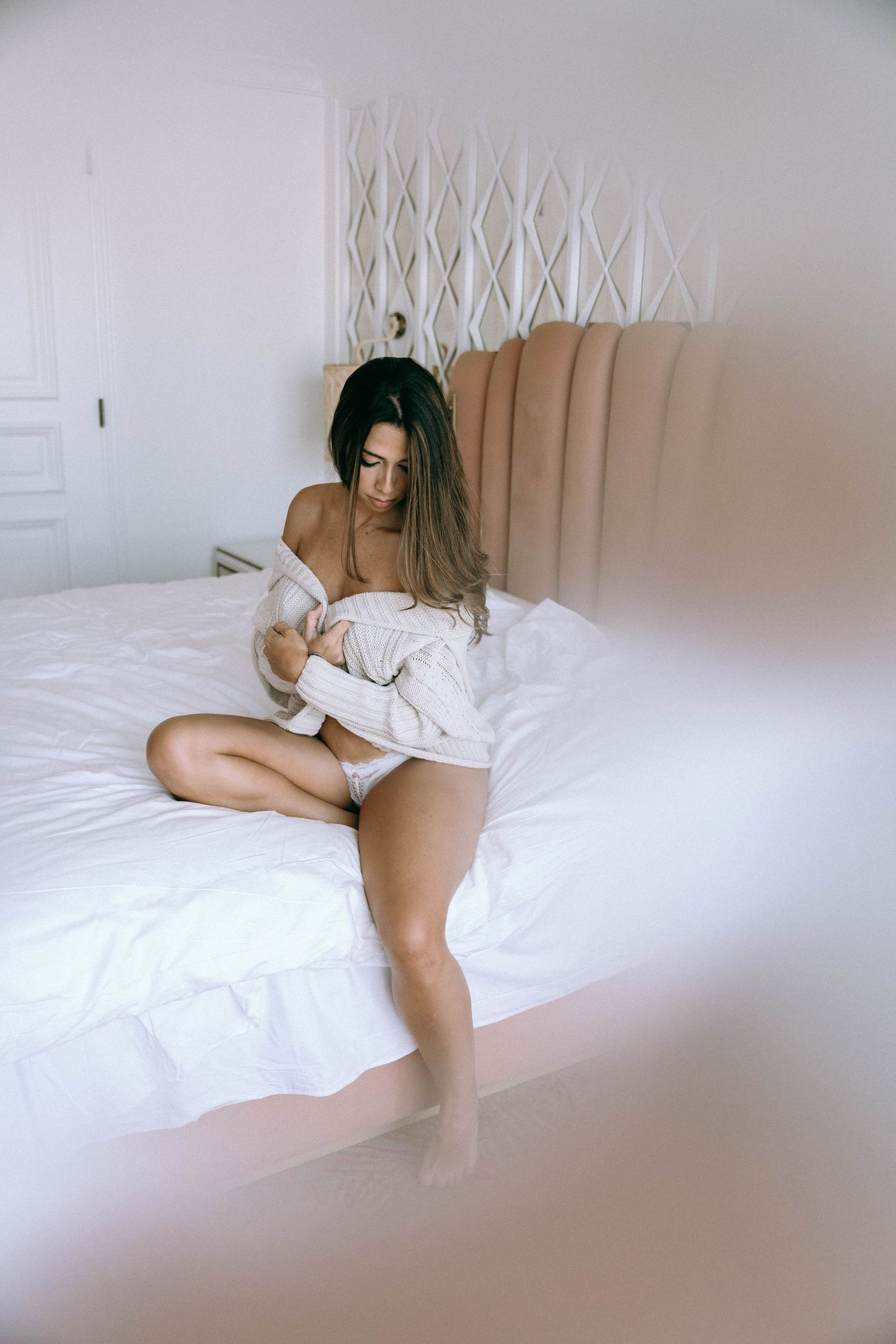 Woman in white underwear and open shirt sitting on a bed with white bedding in a bright bedroom.  Boudoir Palm Beach, FL