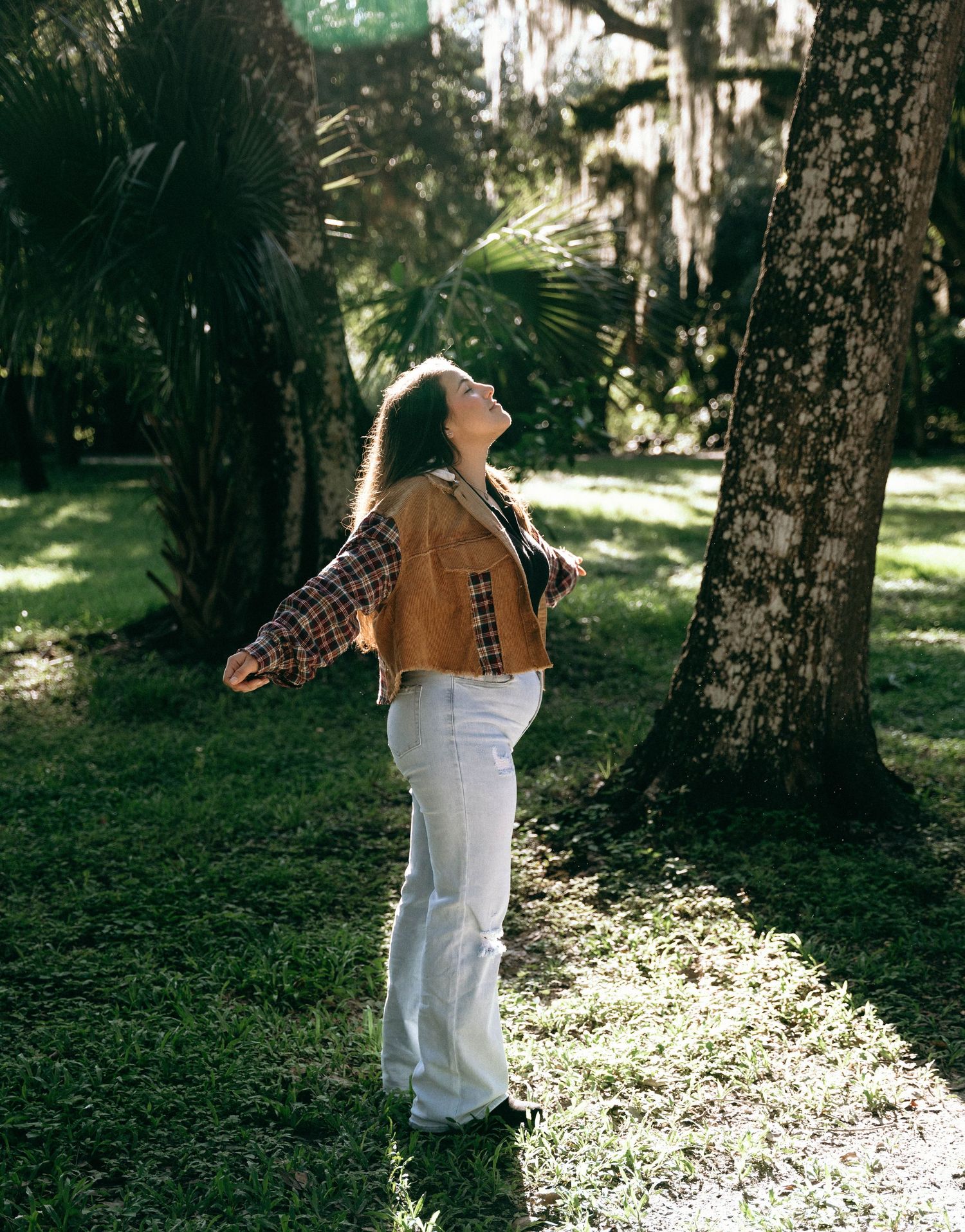 Person in brown jacket and white pants dances joyfully among moss-covered trees.