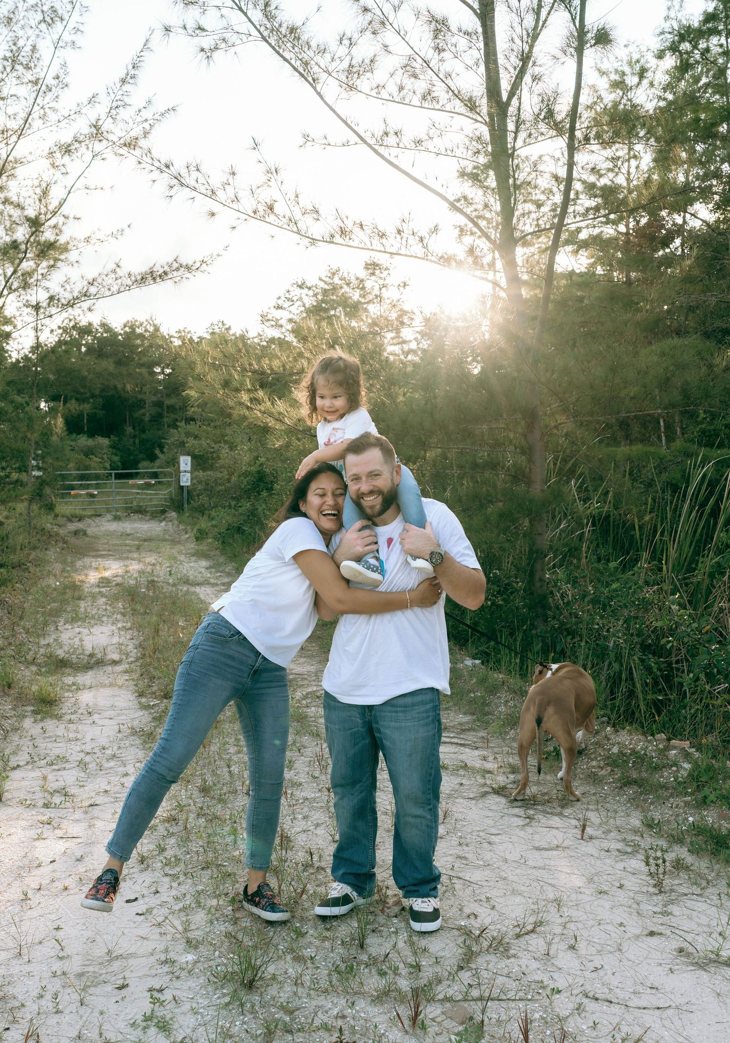 Happy family with toddler on dad's shoulders, mom hugging, and a dog playing on a sandy trail at sunset.