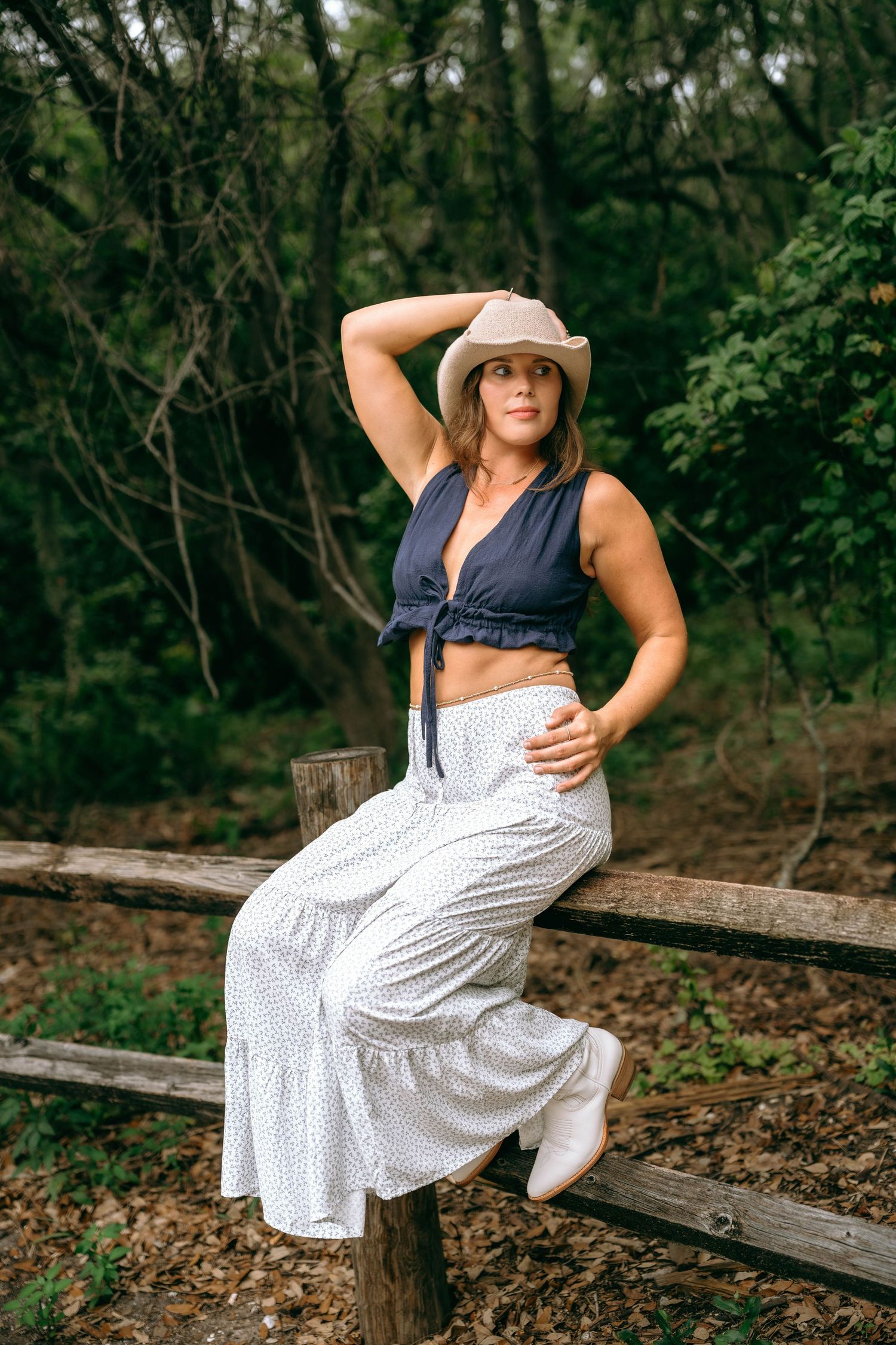 Woman in navy crop top, white wide-leg pants, and cowboy hat posing on a rustic wooden fence in a forest.