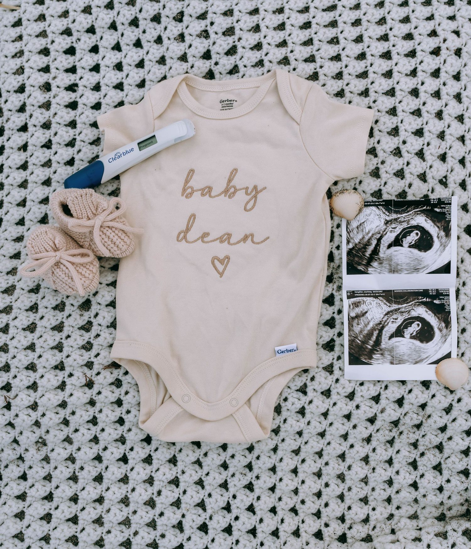 Cream baby onesie reading 'baby dean' with heart, ultrasound photos, booties, and pregnancy test on knit blanket.