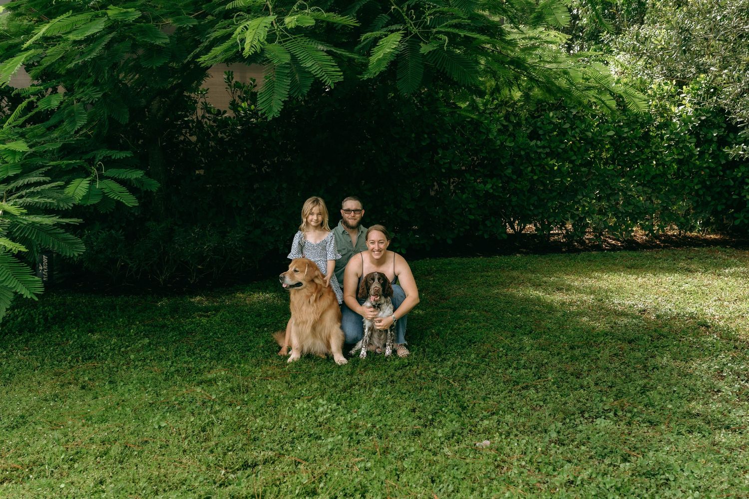 Family with two dogs posing on green grass under large trees in a sunny backyard.