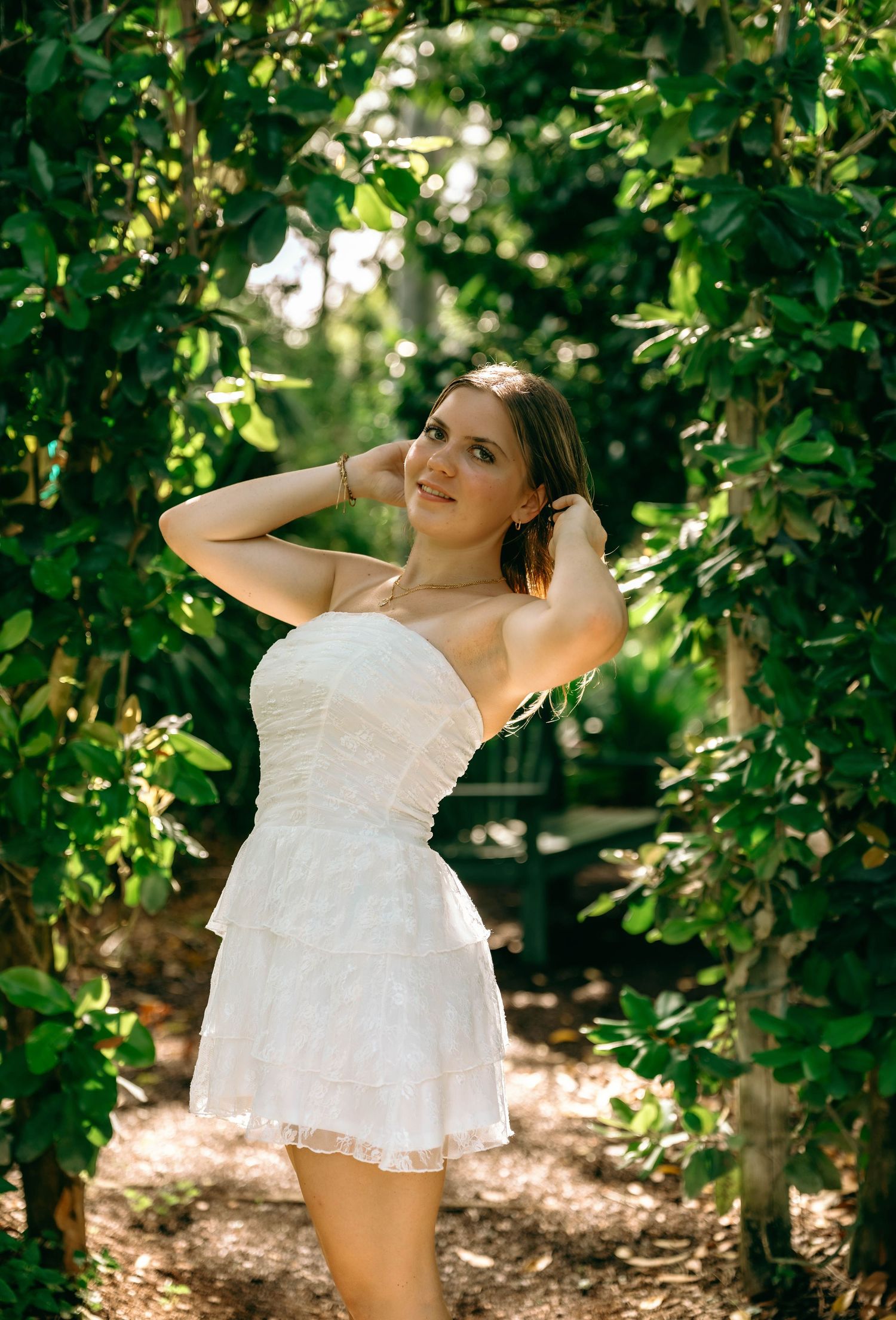 Young woman in white strapless mini dress posing gracefully in a lush green garden archway on a sunny day.
