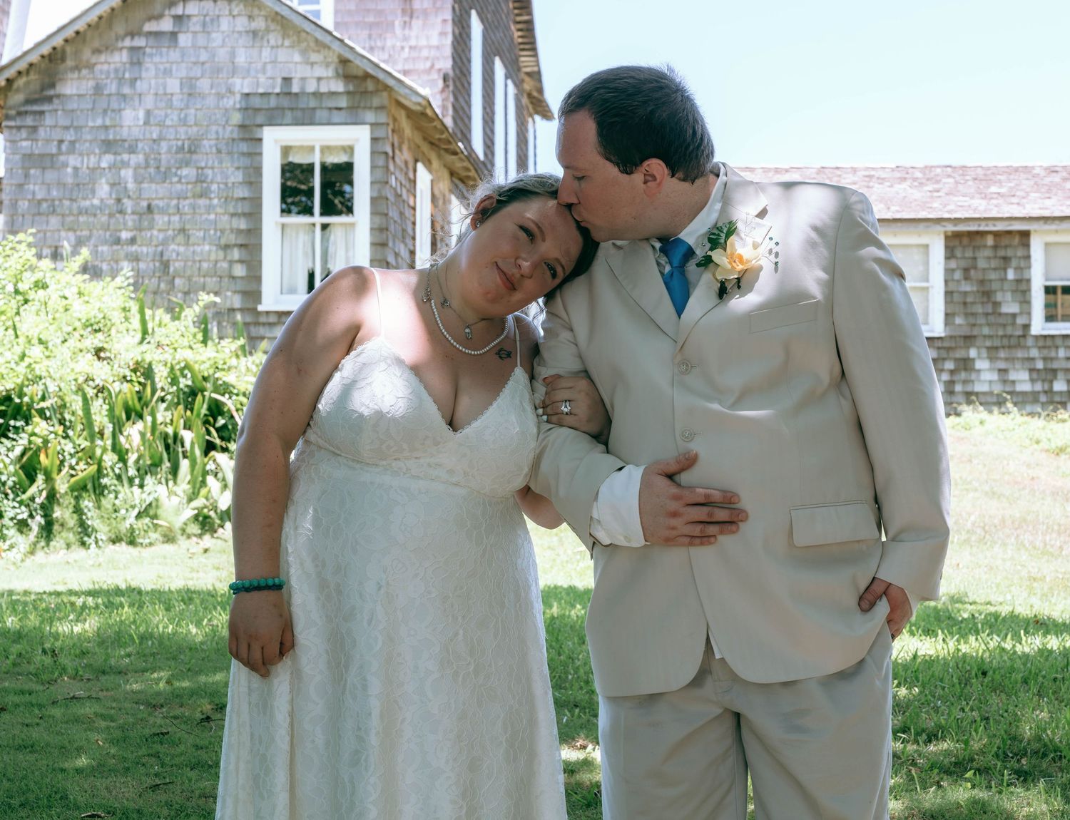Bride in white lace gown leans on groom in tan suit as he kisses her head outdoors near a rustic building.