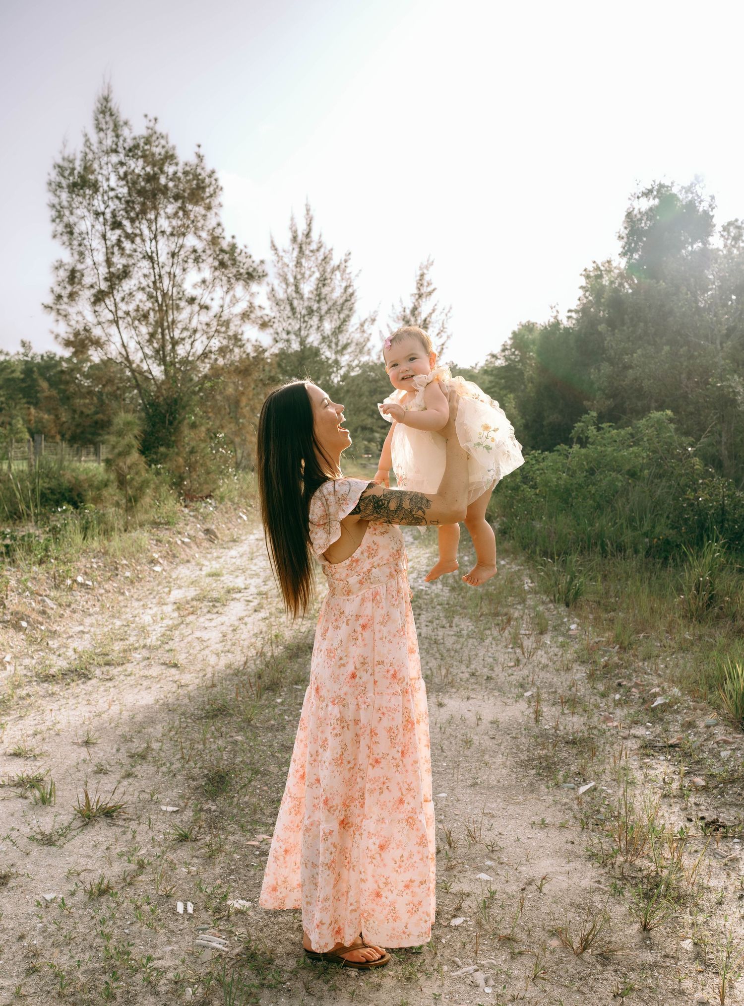 A mother in a floral maxi dress lifts her smiling baby outdoors on a sunny sandy path surrounded by green trees.