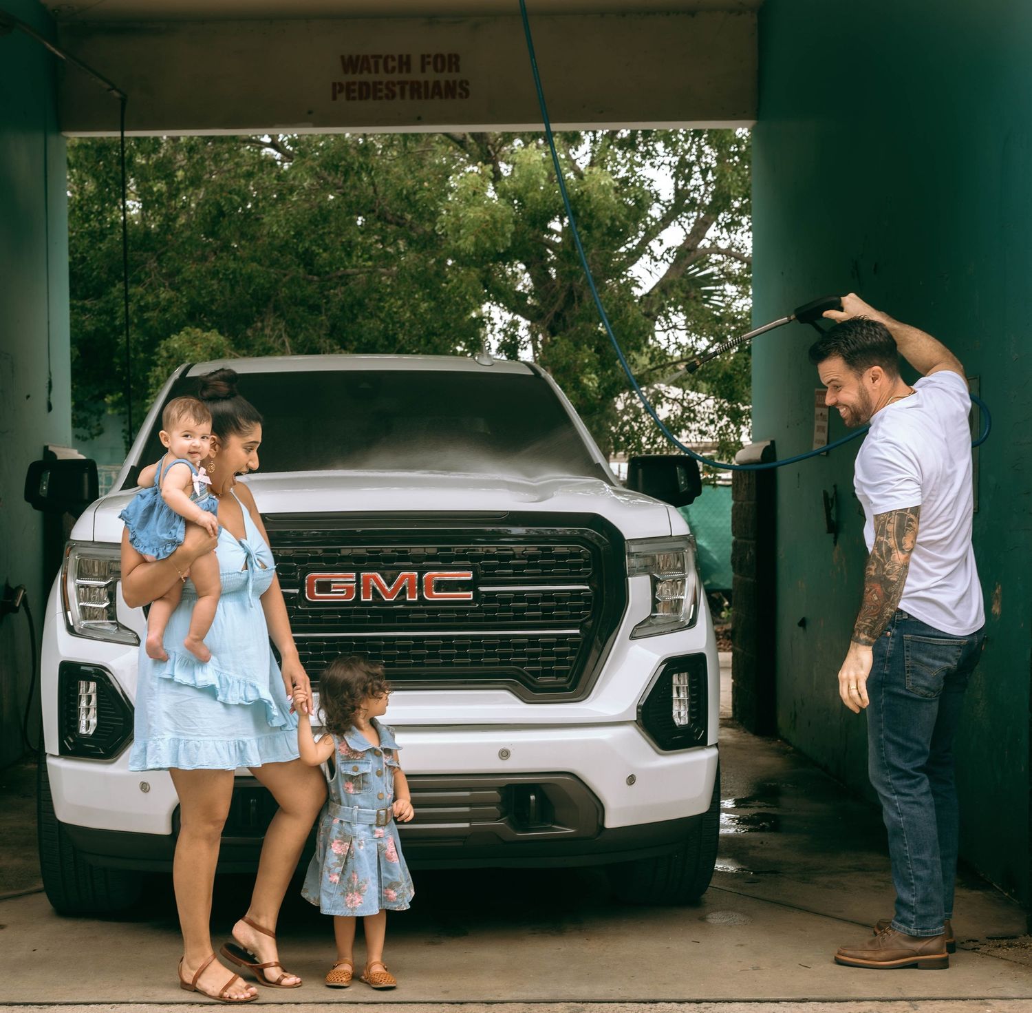 Family posing in front of a white GMC truck at a car wash, with a tattooed man, woman holding baby, and young girl.