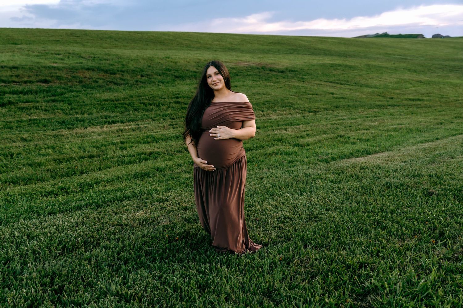 Pregnant woman in brown off-shoulder maxi dress standing in lush green field during maternity photoshoot.