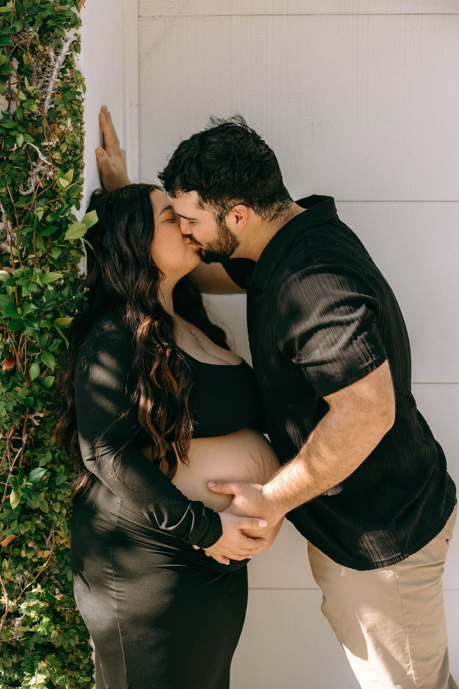 Couple sharing a kiss during maternity photo shoot, man cradling pregnant belly against white wall with ivy. Worth Avenue, Palm Beach, FL