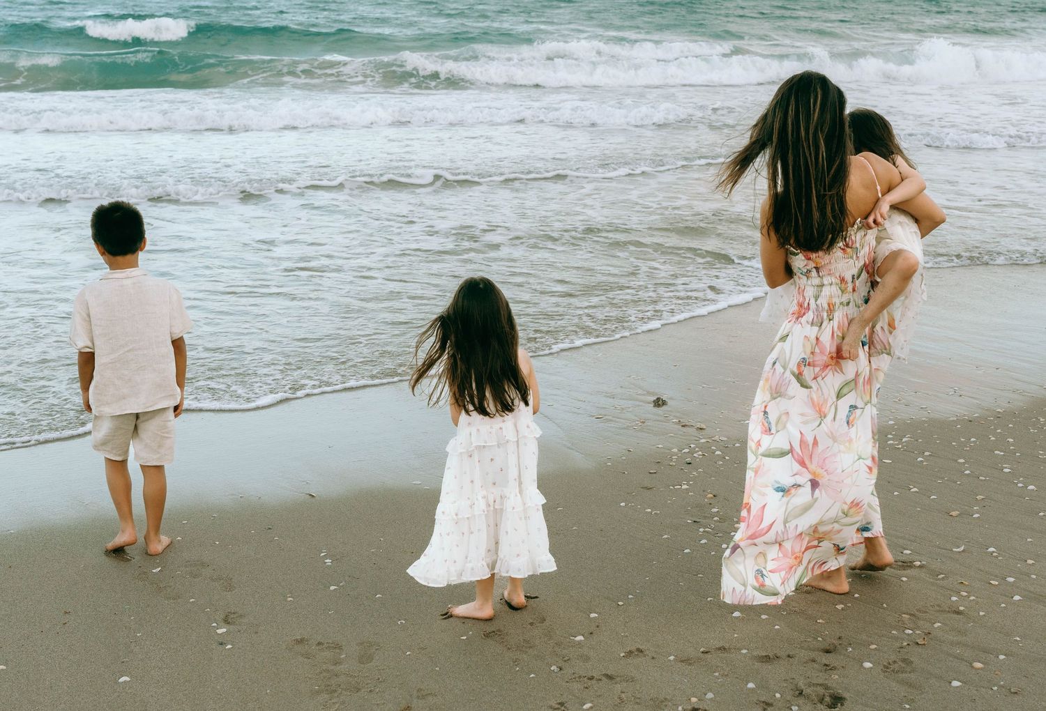 Family of four walking barefoot on a sandy beach shore, mother carrying child in floral dress. Worth Avenue, Palm Beach, FL