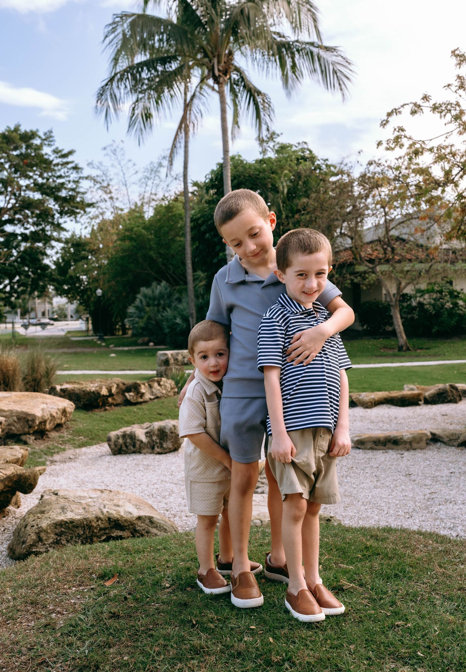 Three young boys pose together outdoors near palm trees and rocks, wearing casual summer outfits.  Family photos at Bradley Park Palm Beach,