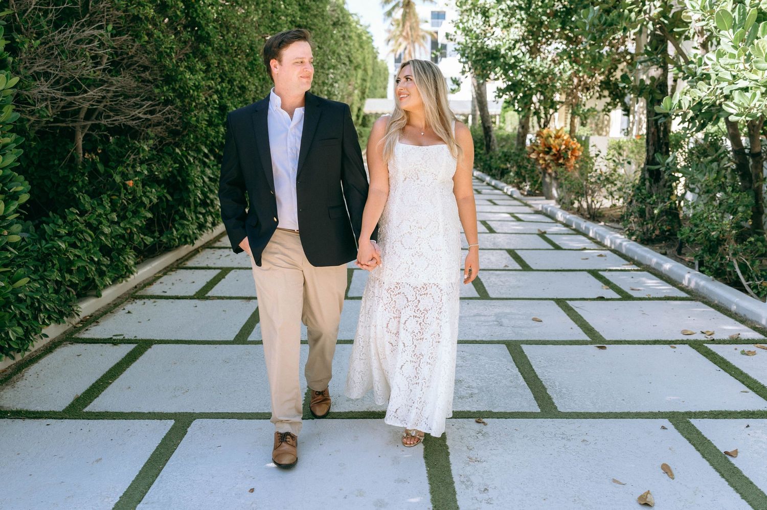 Couple holding hands walking on a stone pathway surrounded by lush greenery, man in navy blazer, woman in white lace dress.