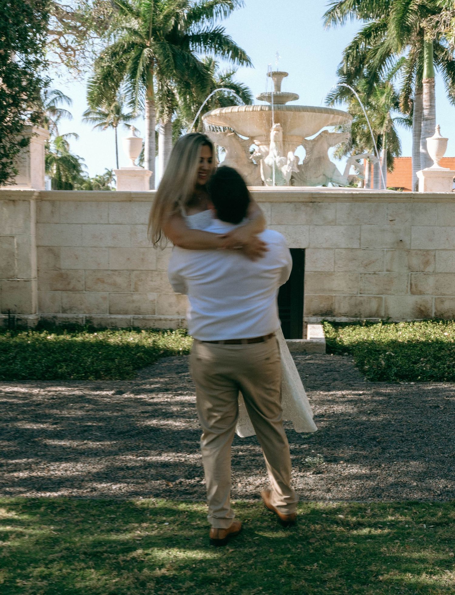 Couple embracing joyfully in a tropical garden with a decorative fountain and palm trees.