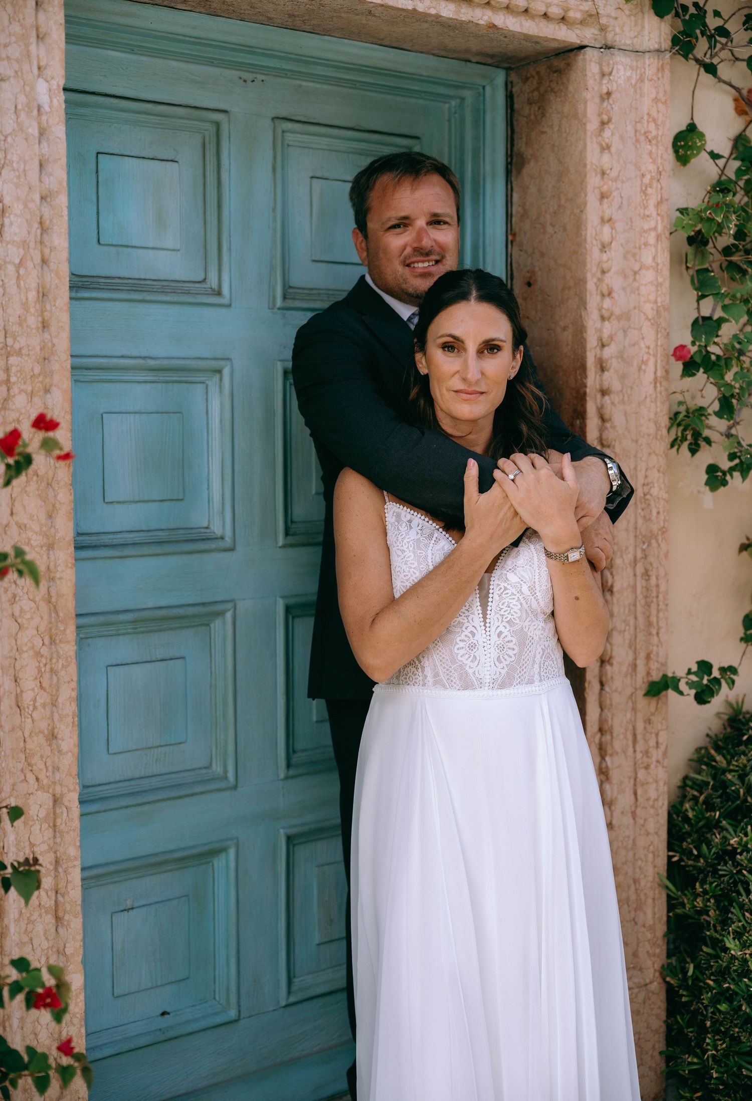 Bride in white lace gown and groom in dark suit embrace in front of a rustic turquoise door with red flowers.