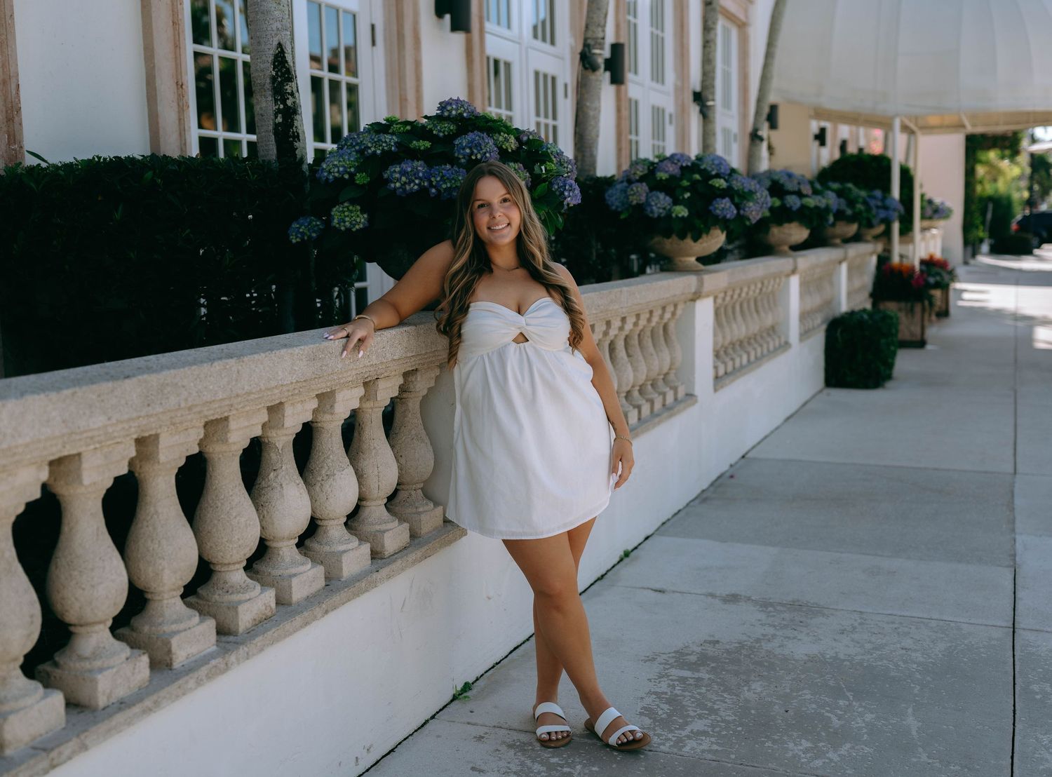 Young woman in white mini dress posing by stone balustrade with hydrangeas.