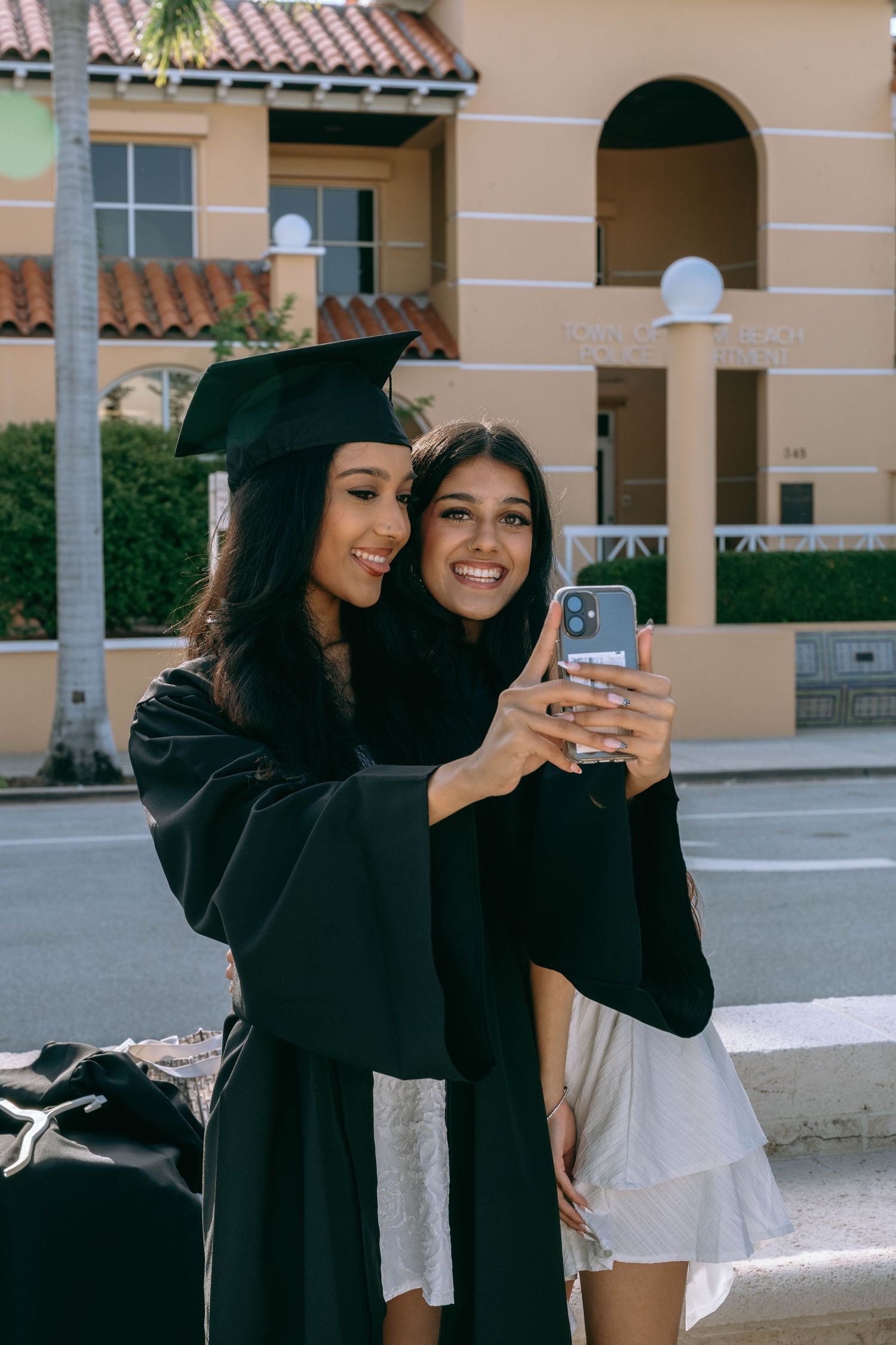 Two young women in graduation attire taking a selfie outdoors near a yellow building on Worth Avenue Palm beach.