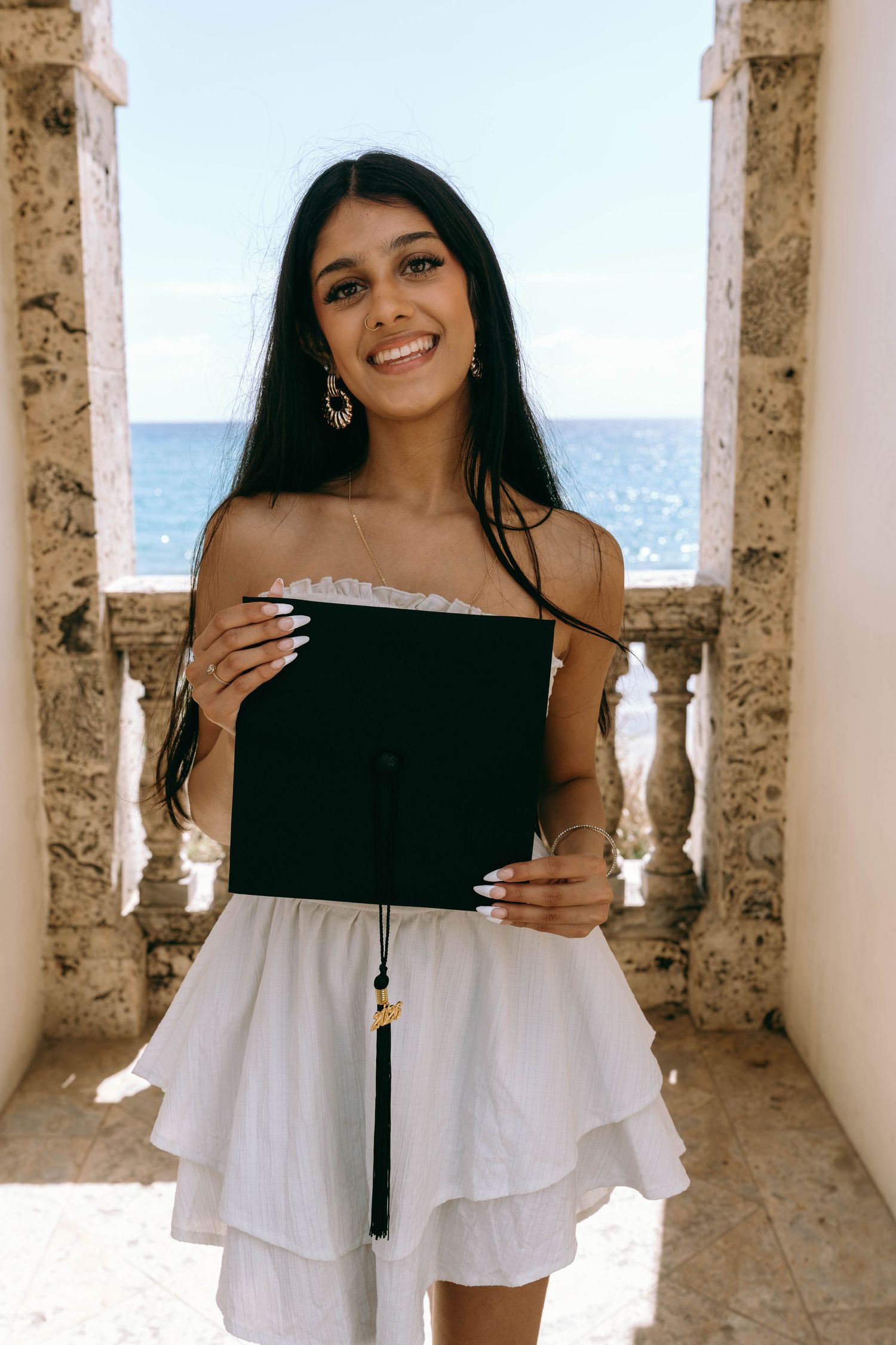 Smiling young woman holding graduation cap with ocean view through stone archway behind her on Worth Avenue Palm beach.