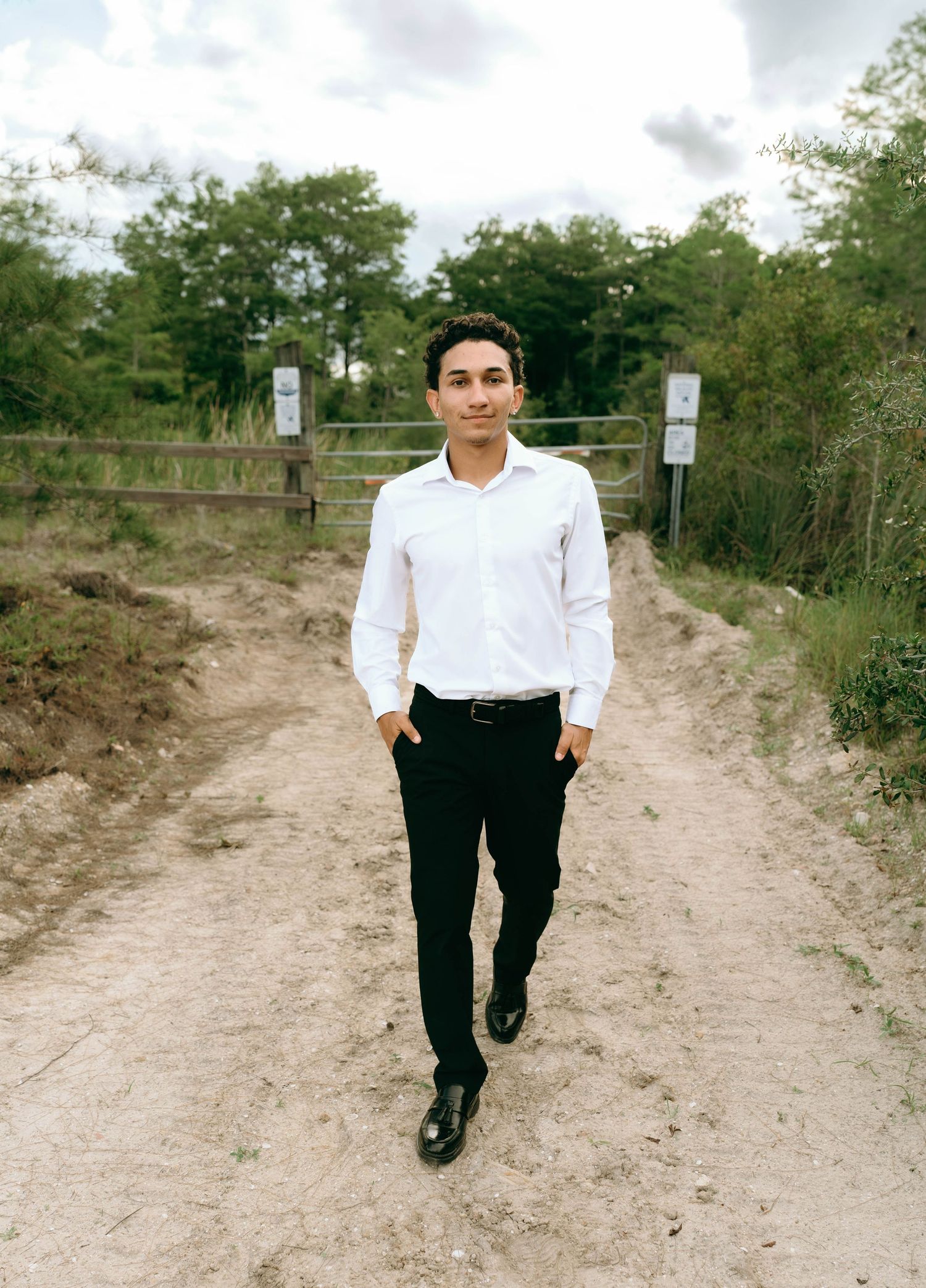 Young man in white dress shirt and black pants walking on a dirt path with trees and gate in background. Senior Photos at Colt Farms