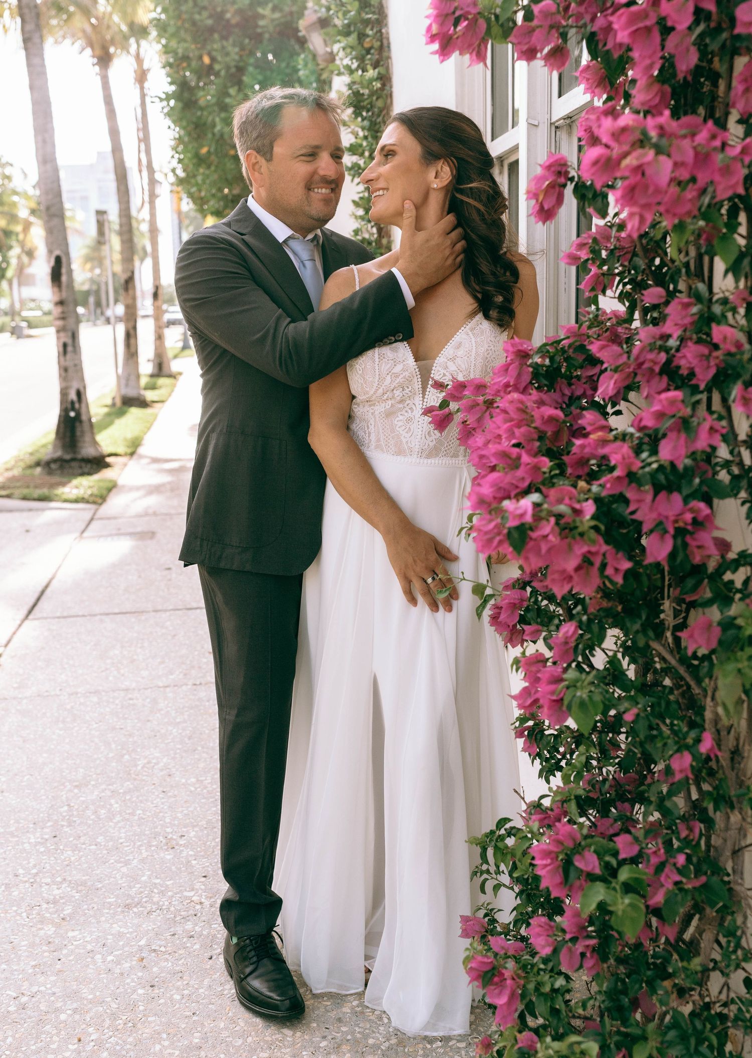 A couple in wedding attire embraces near pink bougainvillea blooms on a sunny sidewalk. Worth Avenue, Palm Beach, FL