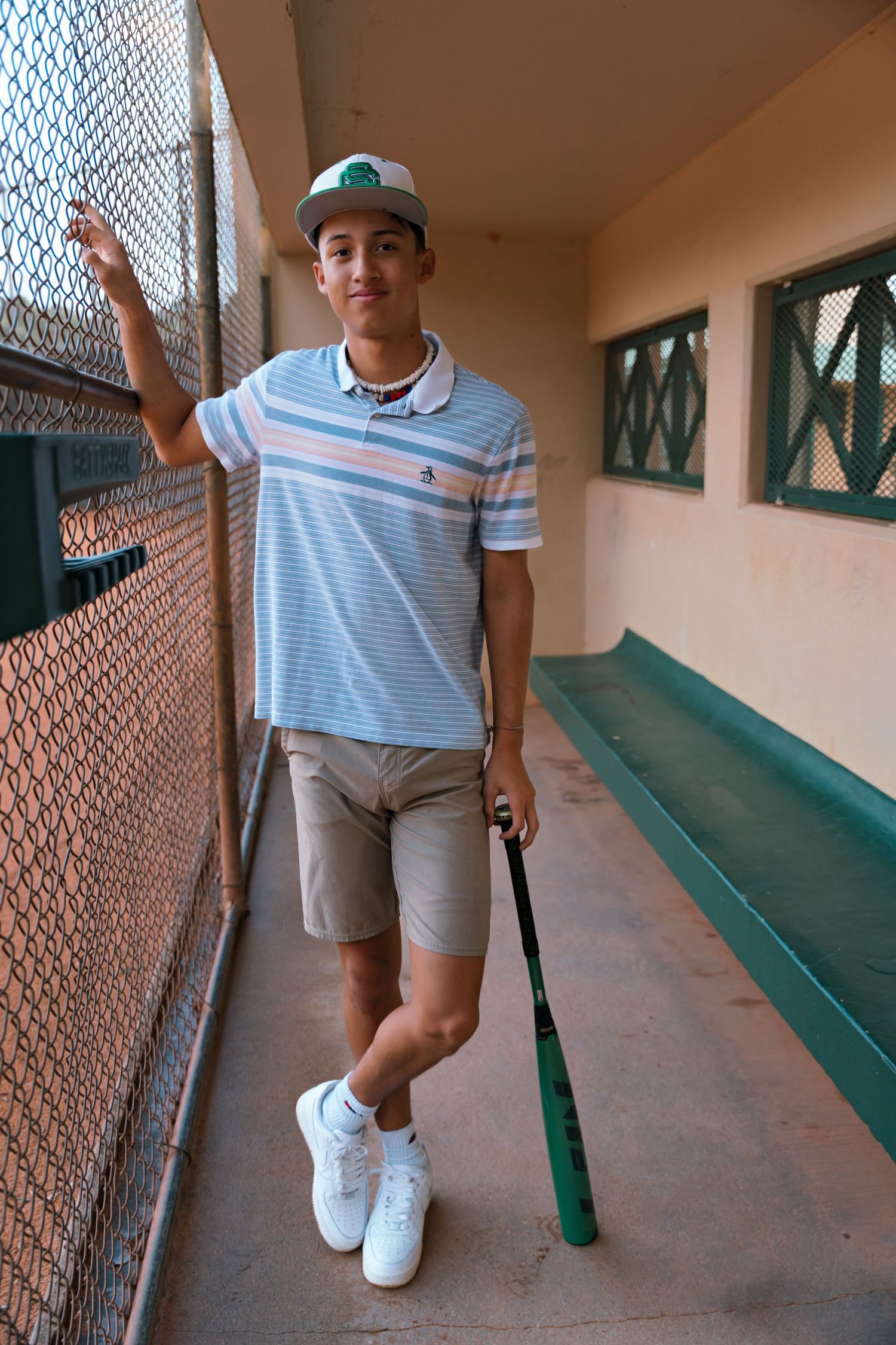 Baseball player in striped shirt and shorts standing in a dugout with bat and glove.