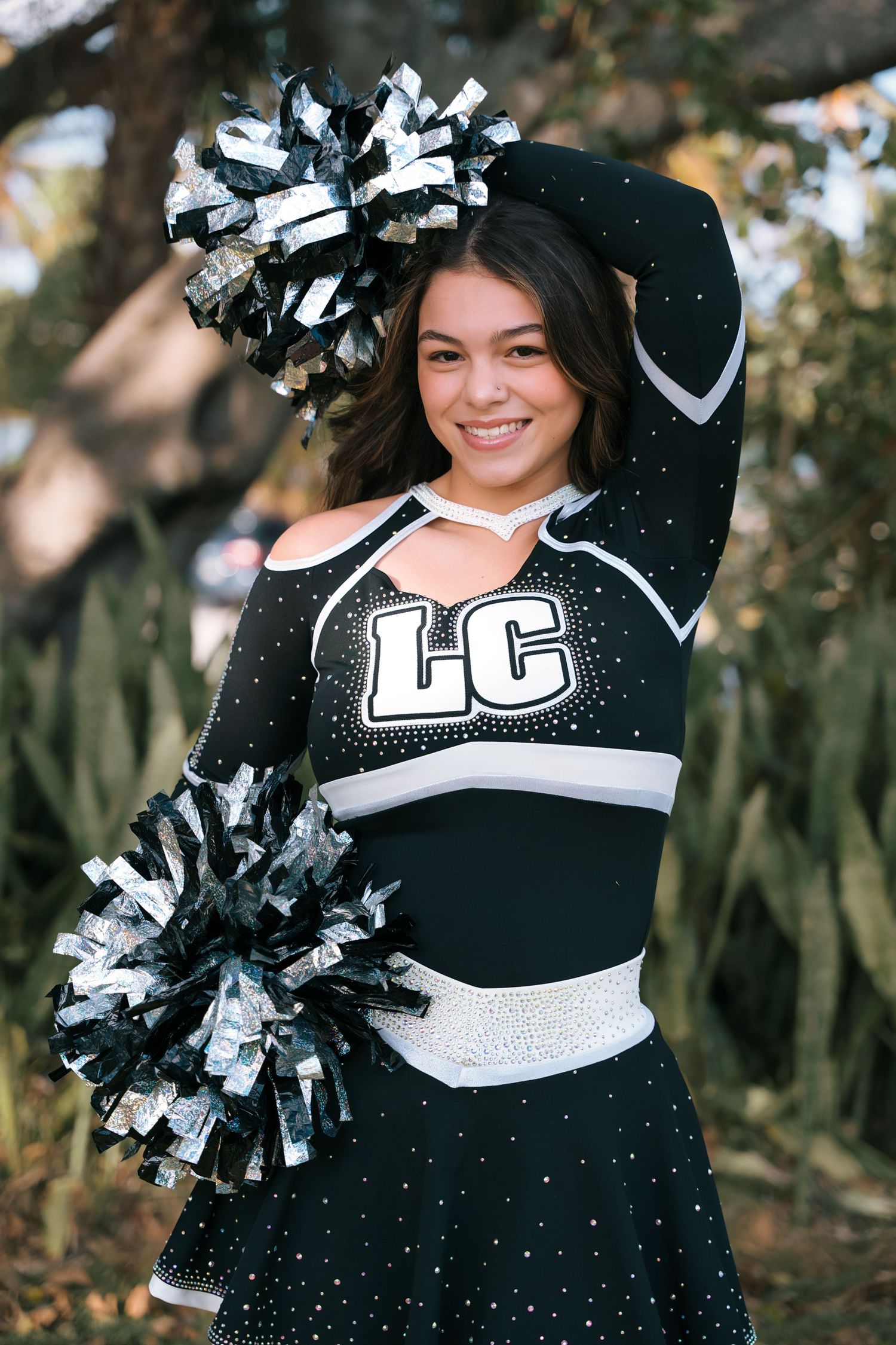 A cheerleader in black and white uniform with pom poms poses outdoors.
