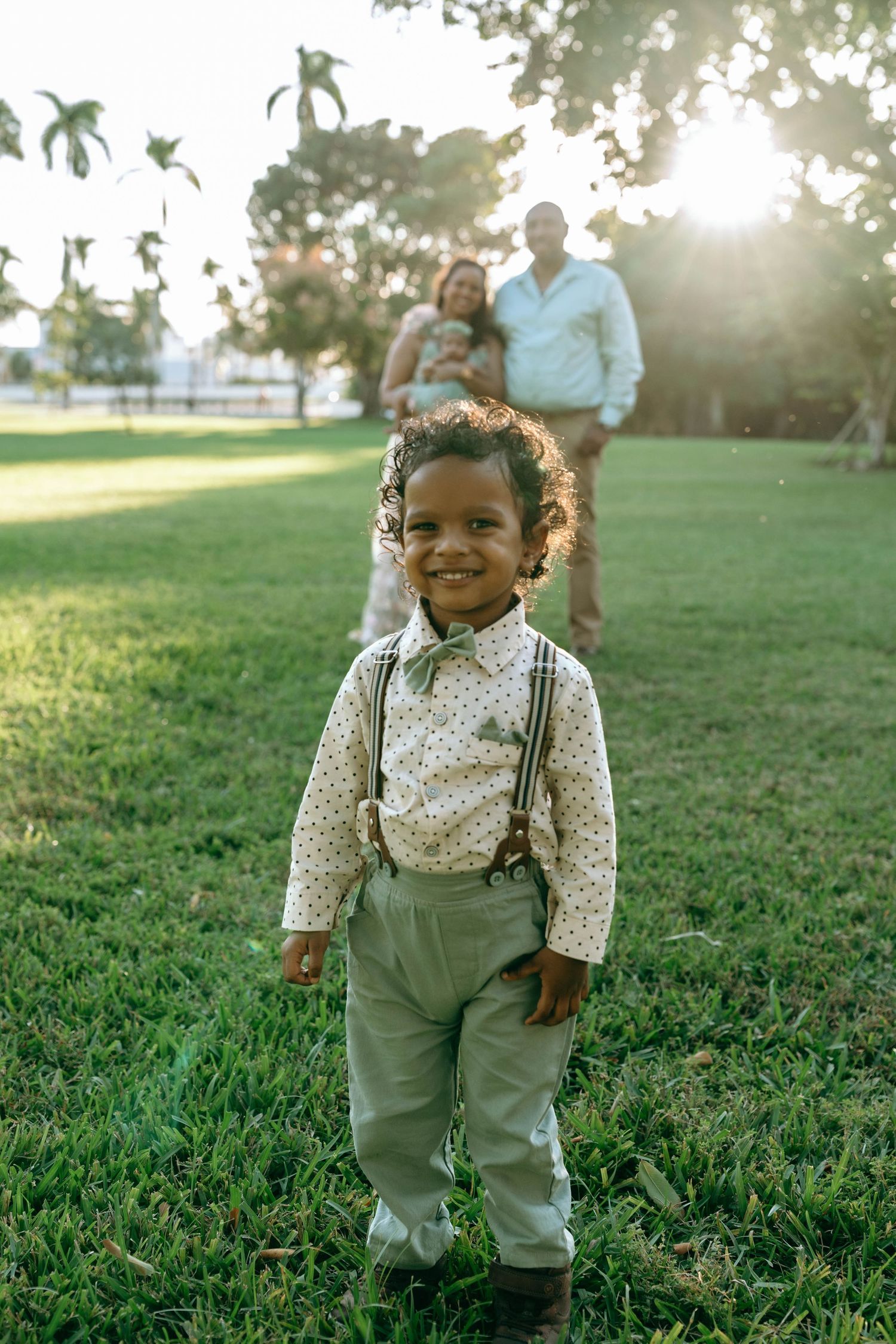 A toddler wearing suspenders and a floral shirt stands on green grass during golden hour.