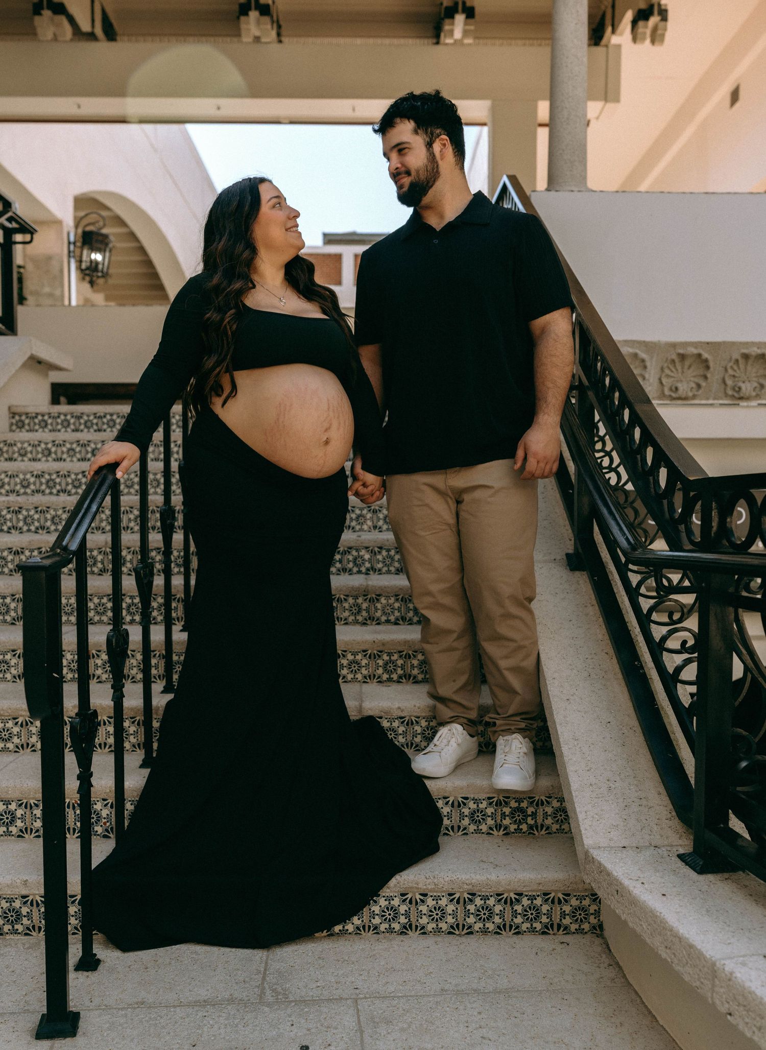 Pregnant woman in black gown holds hands with partner on decorative staircase during maternity photoshoot. Worth Avenue, Palm Beach, FL