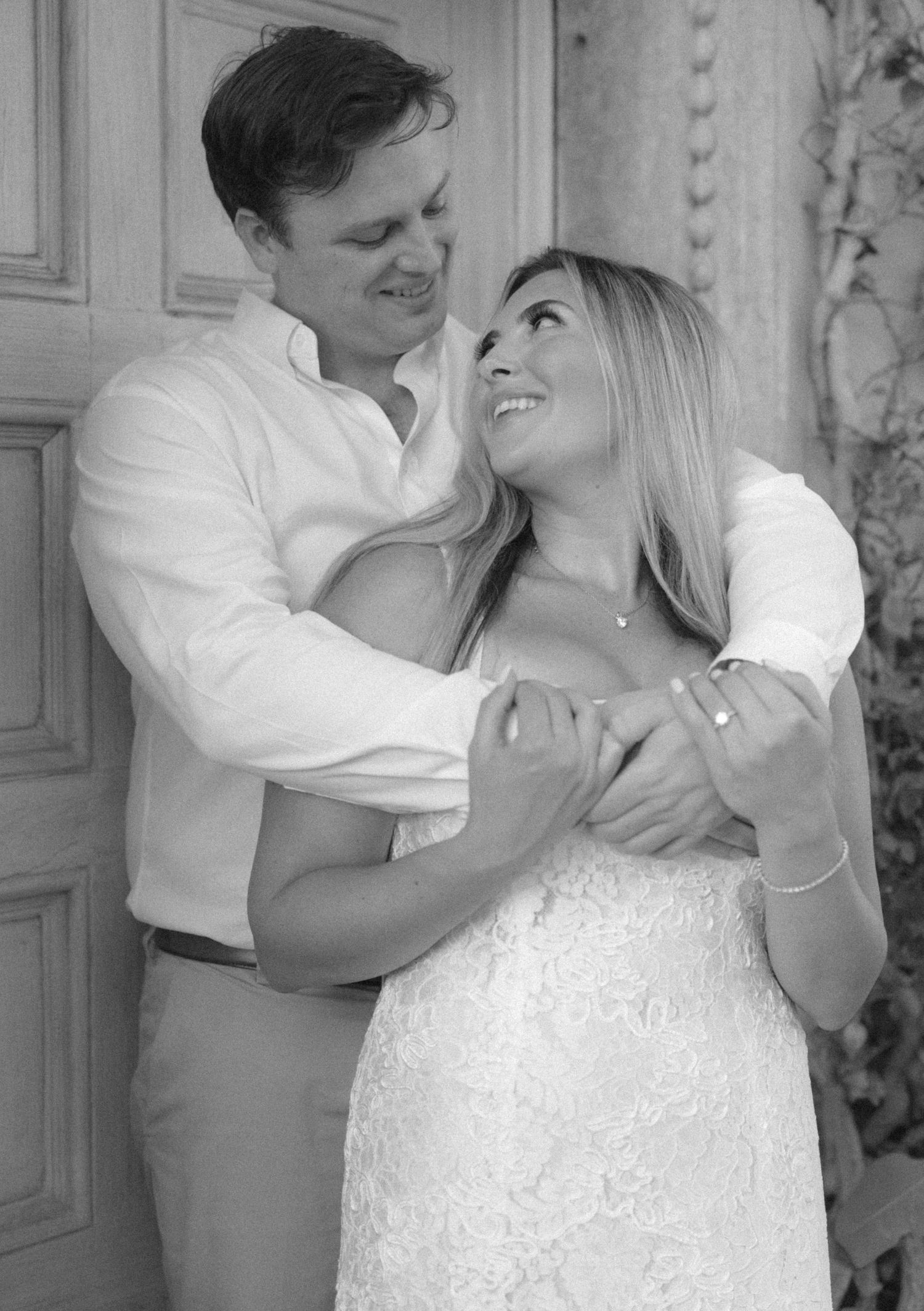 Black and white photo of a smiling couple embracing near a wooden door, woman in lace dress gazing up at man.