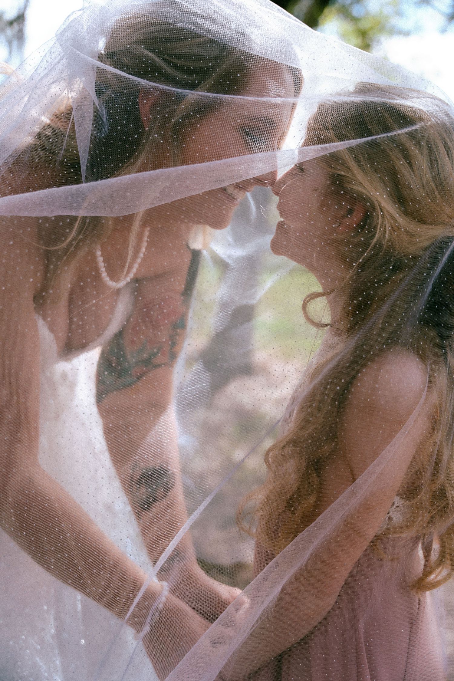 Bride and bridesmaid share an intimate moment under a dotted wedding veil outdoors.