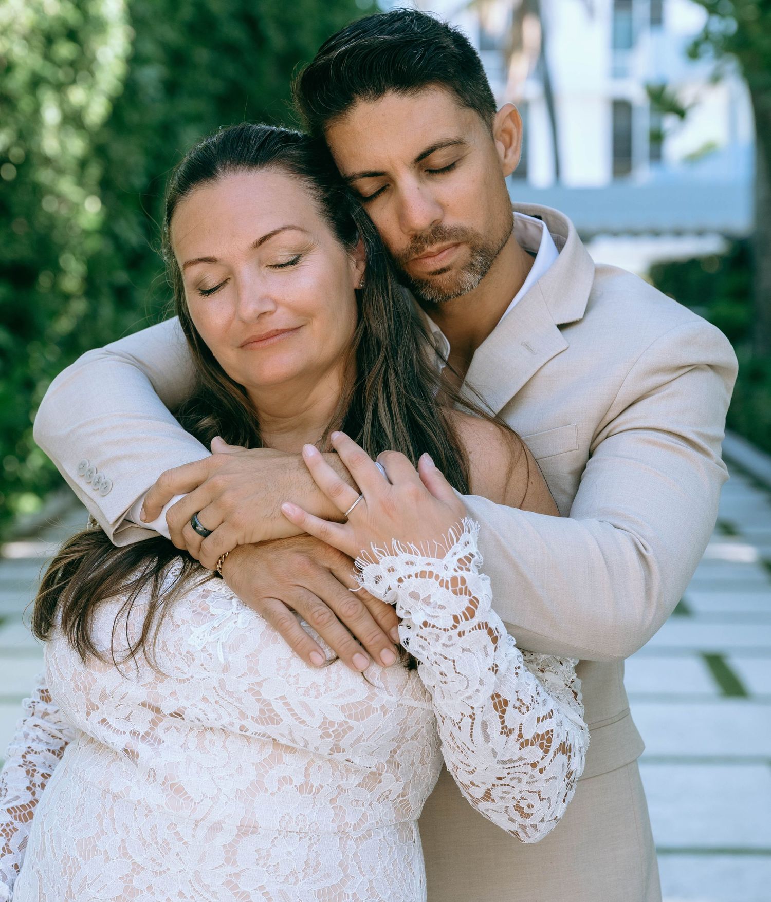 Groom in tan suit tenderly embracing bride in lace dress in garden setting.