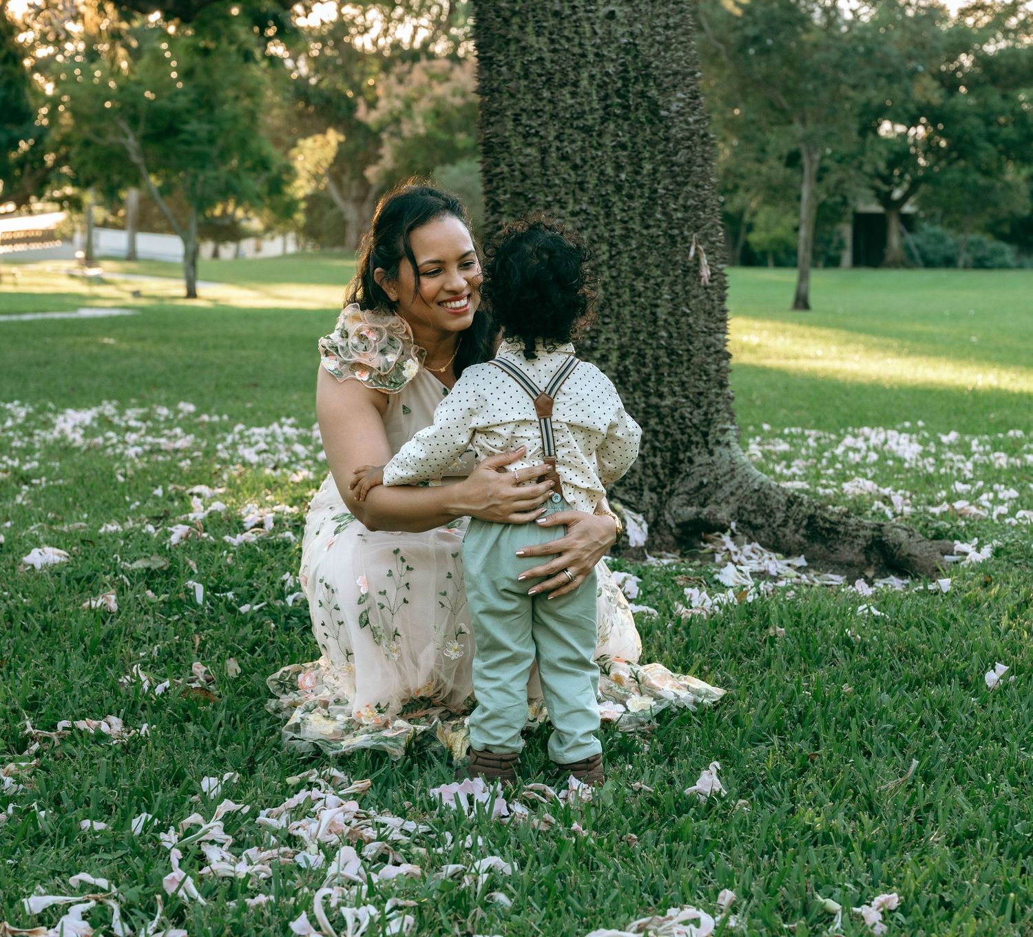 A family sits together on grass surrounded by scattered white flower petals in a sunlit park setting.
