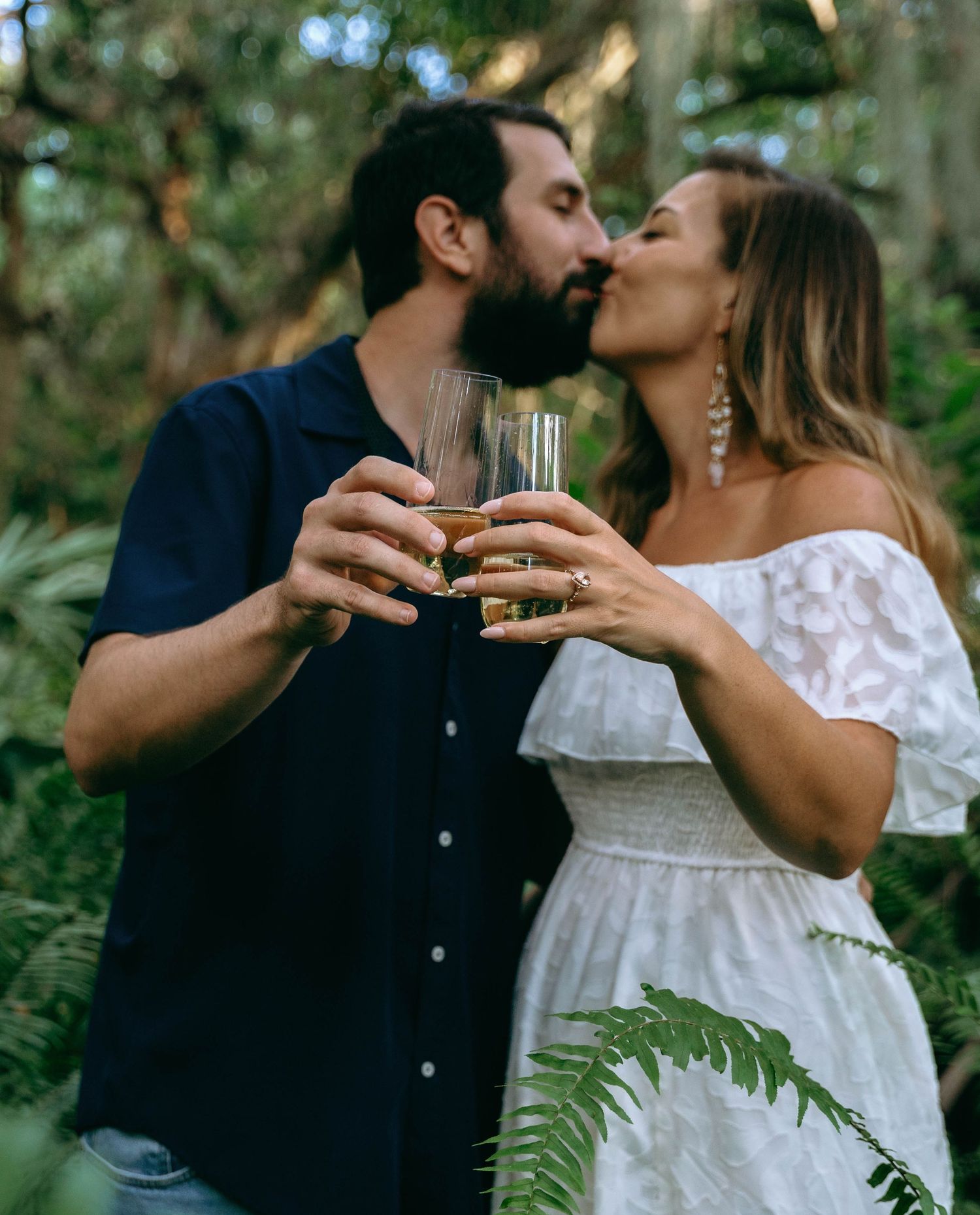 A couple shares champagne glasses while surrounded by lush green foliage.