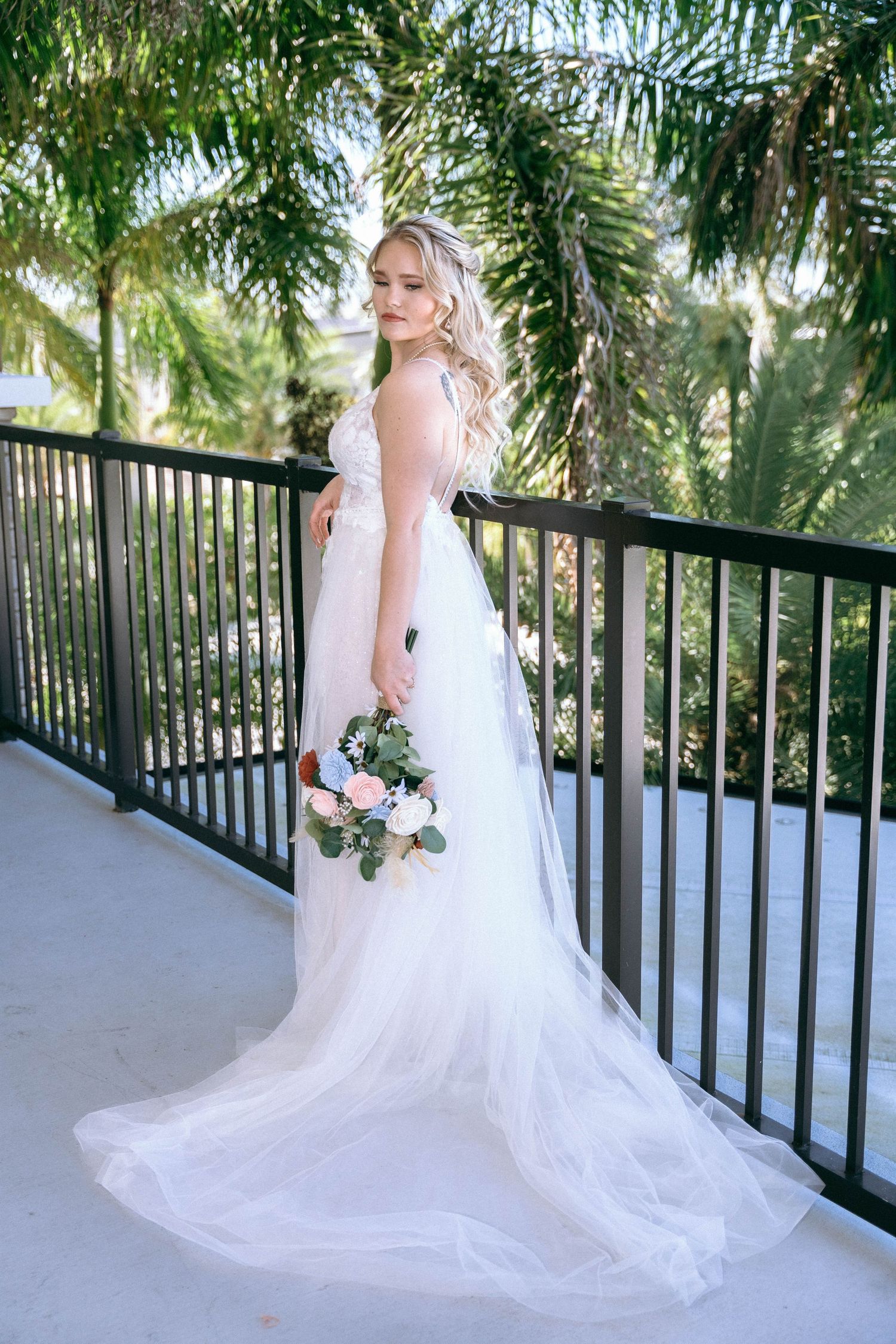 A flowing white wedding dress train spreads across an outdoor balcony with tropical palm trees in view.