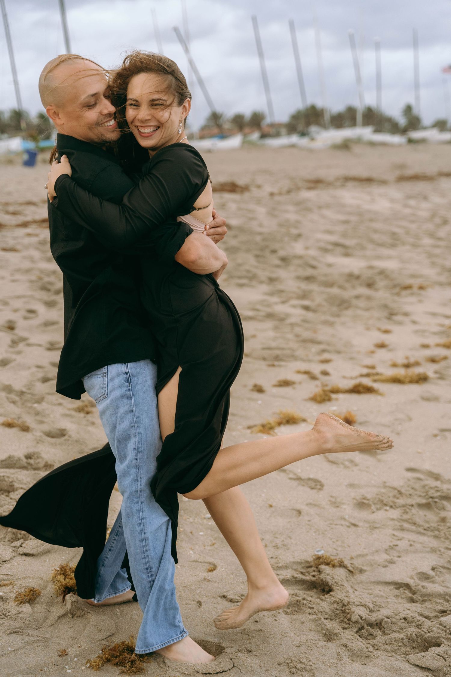 Playful couple embracing and dancing on sandy beach in casual attire.