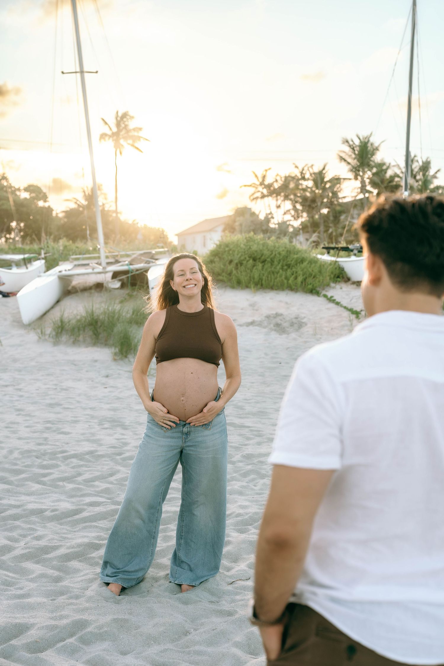 Pregnant woman in brown crop top and jeans smiling on sandy beach at sunset with sailboats and palm trees.