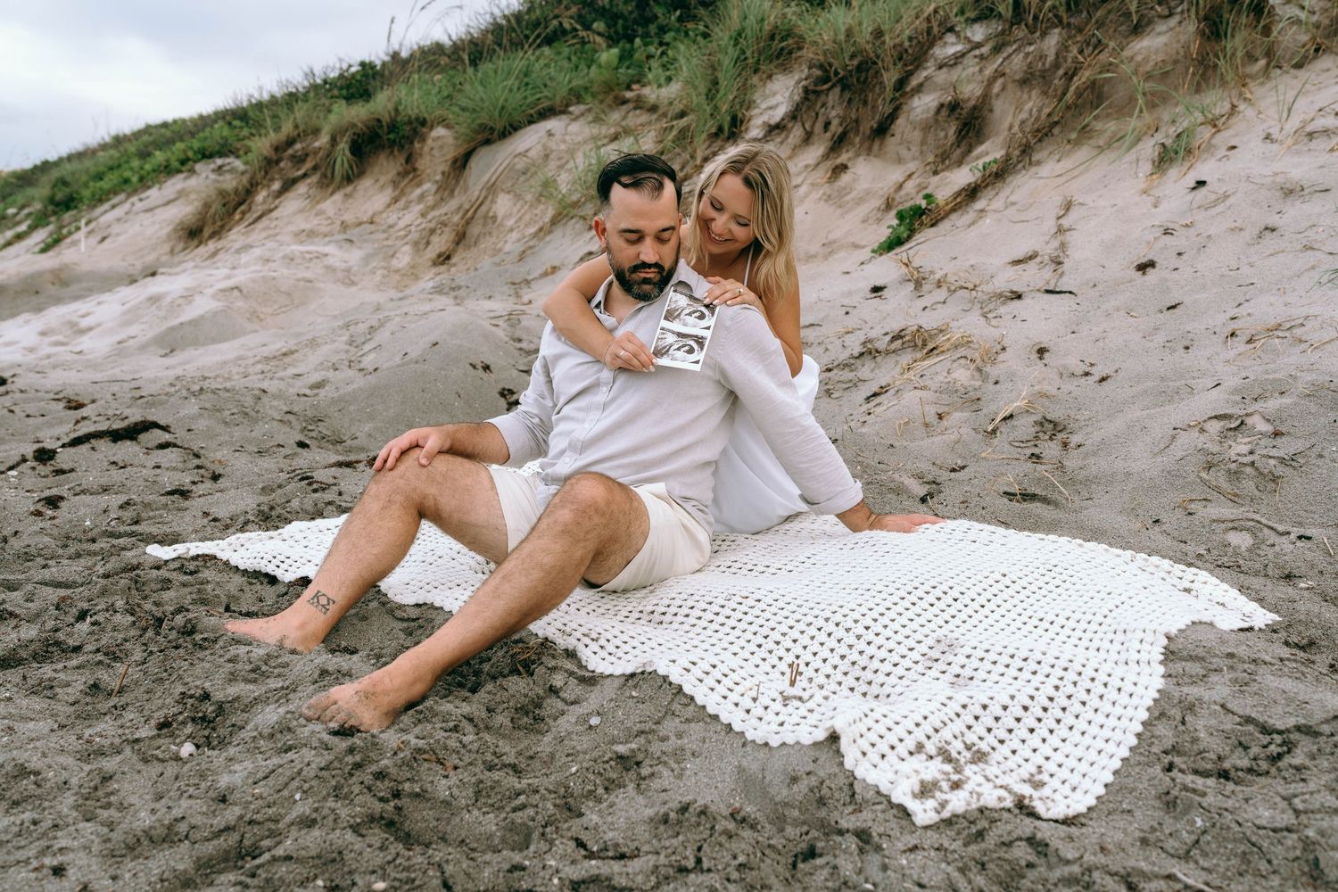 Couple sitting on sandy beach, woman embracing man from behind on white blanket near sand dunes.