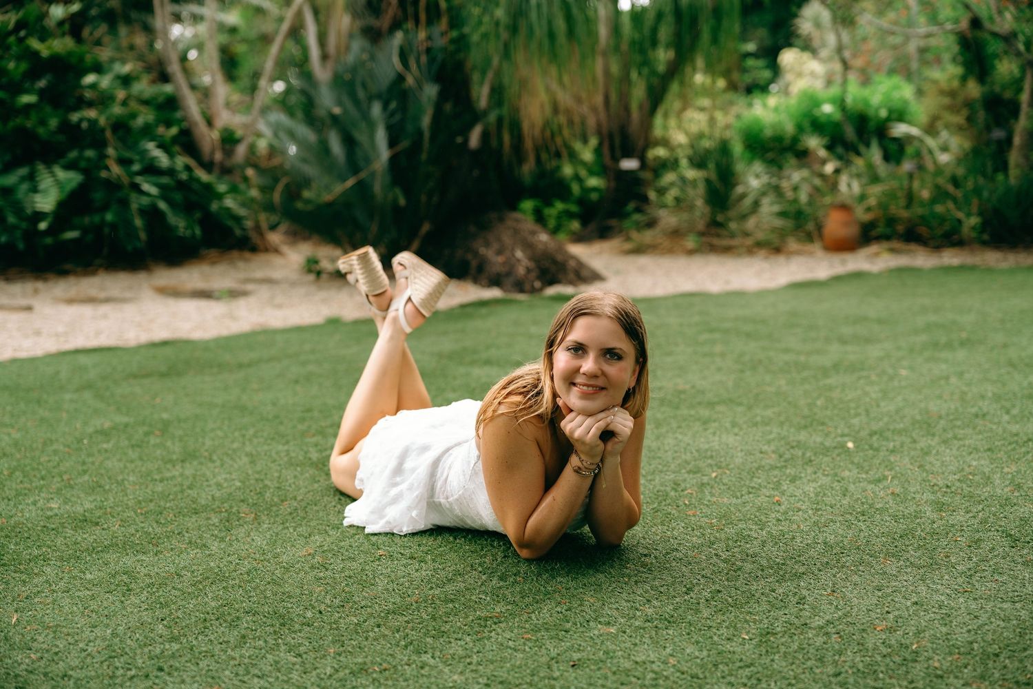 Young woman in white dress lying on green grass lawn smiling with tropical garden background.