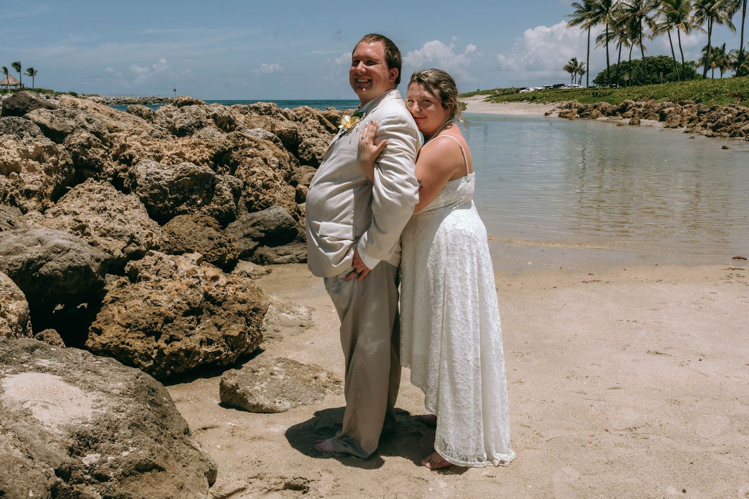 Bride in white dress and groom in tan suit pose together on a rocky beach with palm trees and ocean backdrop.