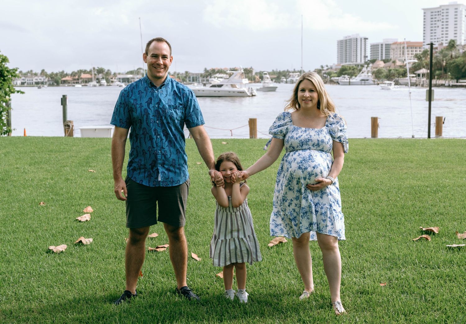 Pregnant woman and man hold hands with young girl on green lawn by marina with boats and waterfront buildings.