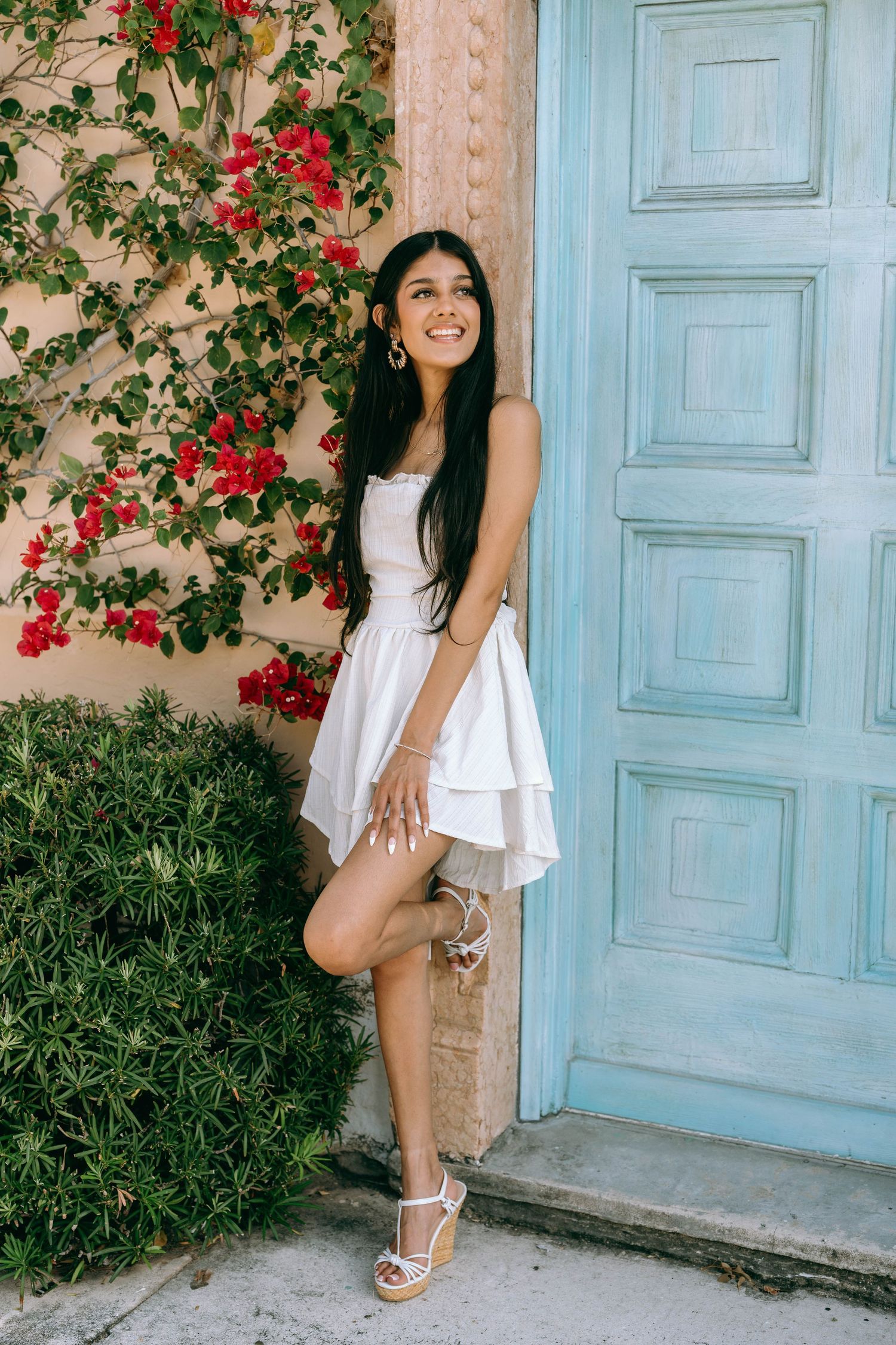 Smiling young woman in white dress posing by blue door with red bougainvillea flowers on Worth Avenue Palm beach.
