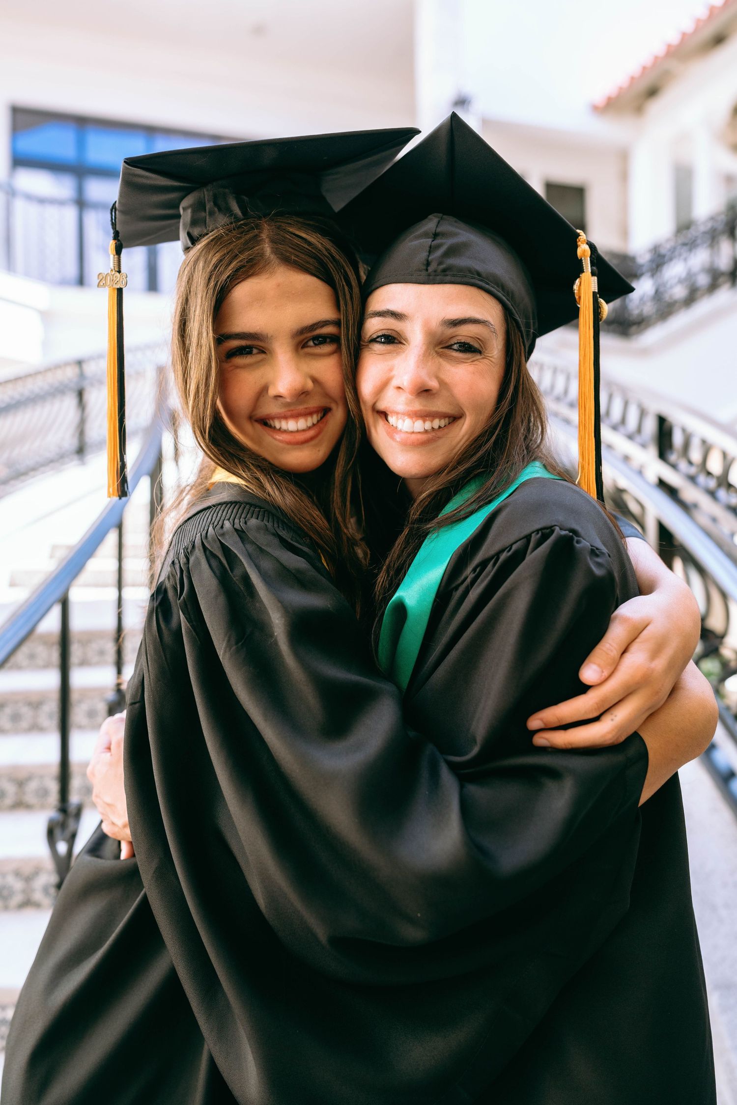 Two smiling female graduates in black caps and gowns hugging at graduation ceremony on Worth Avenue Palm beach.