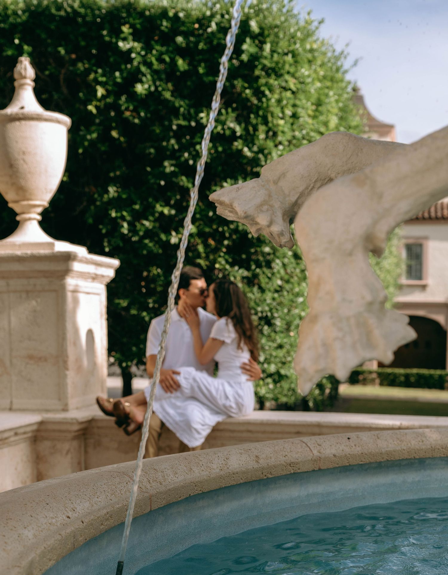 Romantic couple kissing while sitting on the edge of an ornate stone fountain in a lush garden.  Engagement photos on Worth Avenue