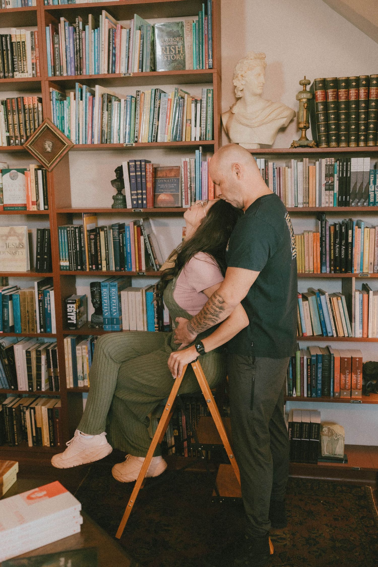 Couple sharing a romantic kiss in a cozy home library with wooden bookshelves filled with books.