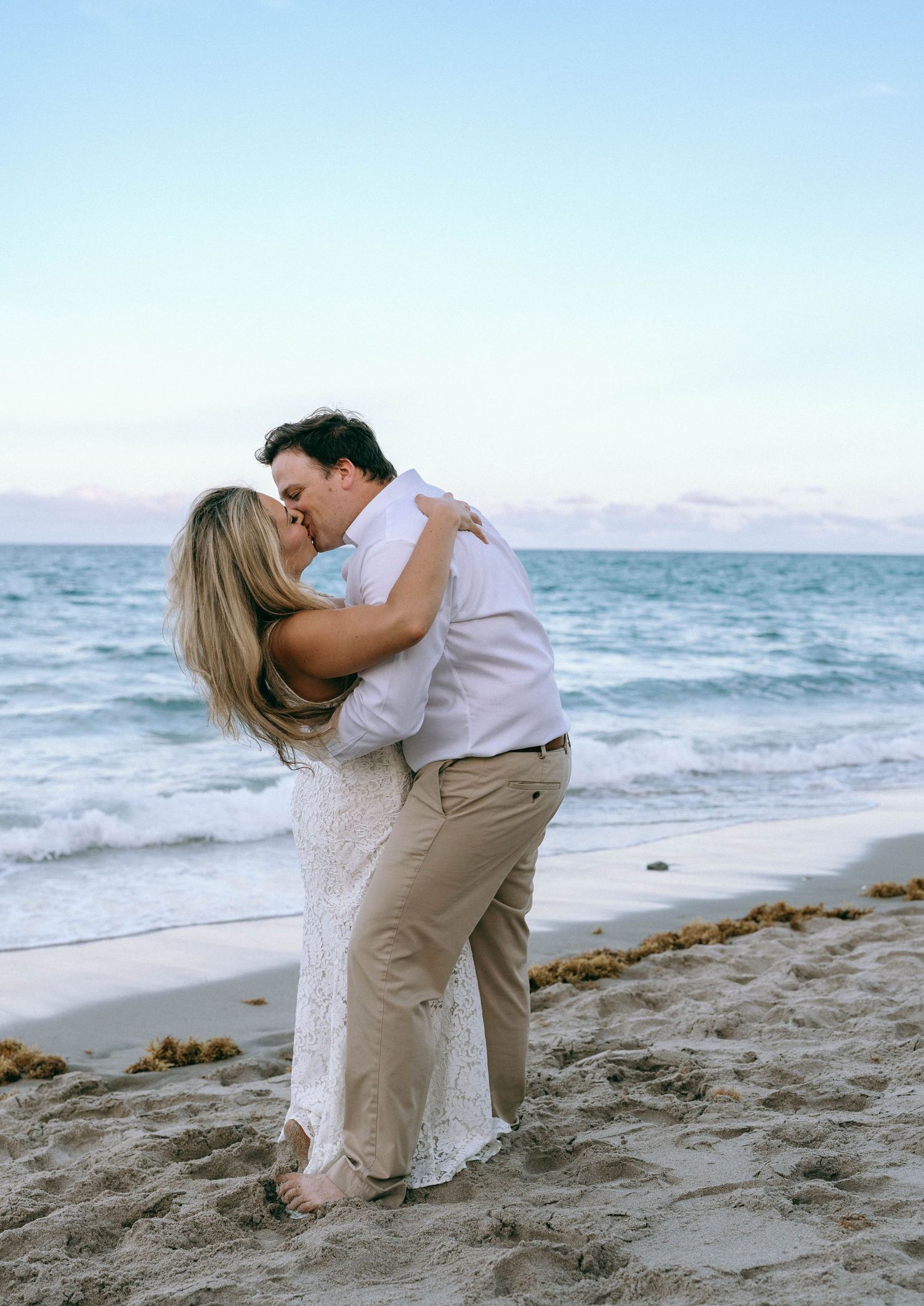Romantic couple sharing a kiss on the beach at sunset, woman in white lace dress embracing man in white shirt.