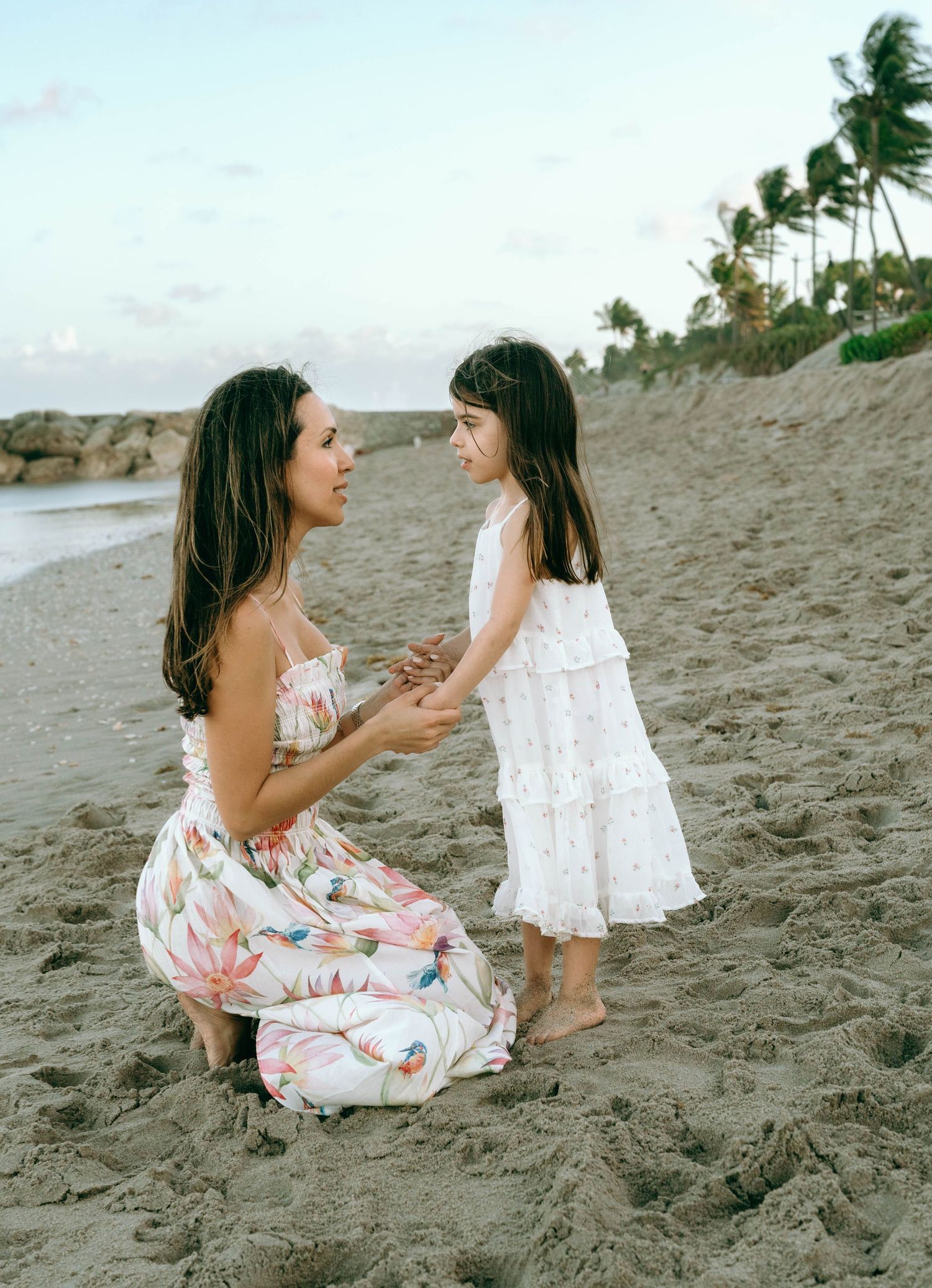 A woman in a floral dress kneels on a sandy beach, holding hands with a young girl in a white dress near palm trees. Worth Avenue